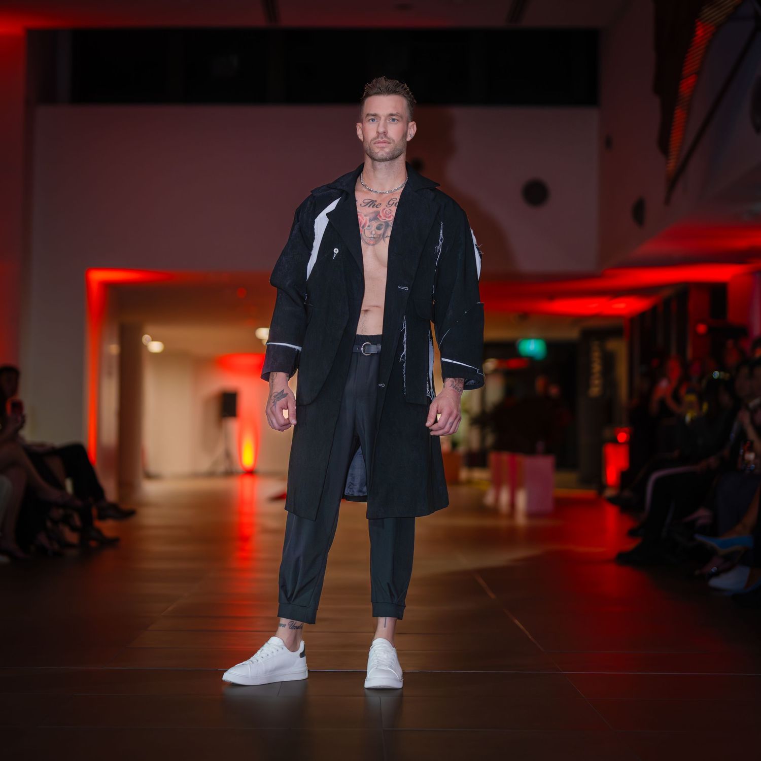 Fashion model showcases dark denim outfit with white sneakers on illuminated catwalk with red ambient lighting.