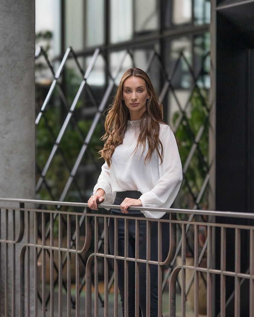 A woman in a white blouse stands on a balcony with a metal railing and lush greenery in the background.