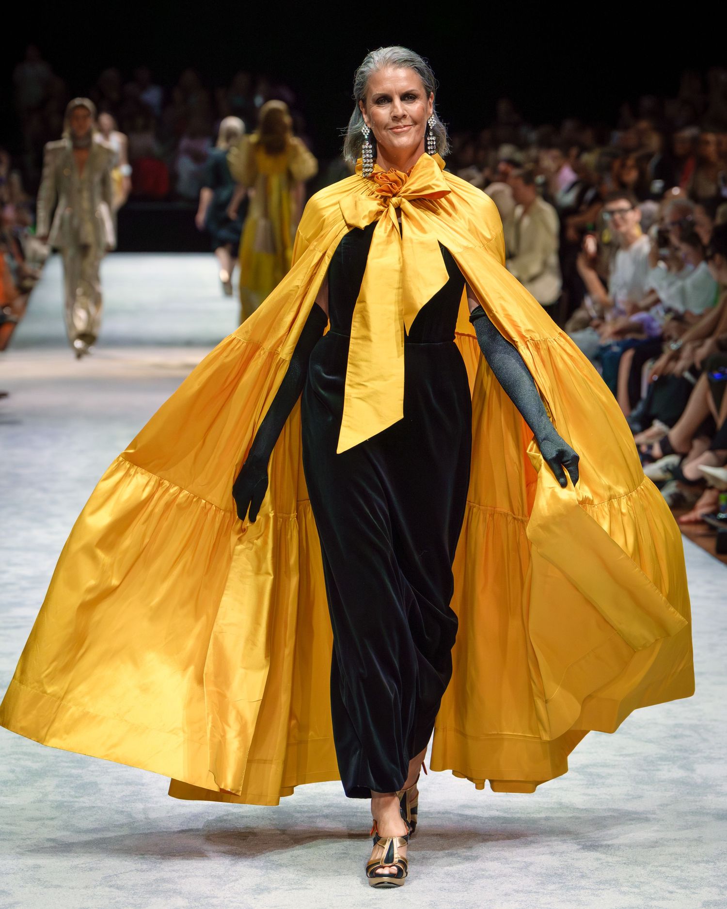Model walks runway in dramatic black dress with flowing yellow cape at fashion show.