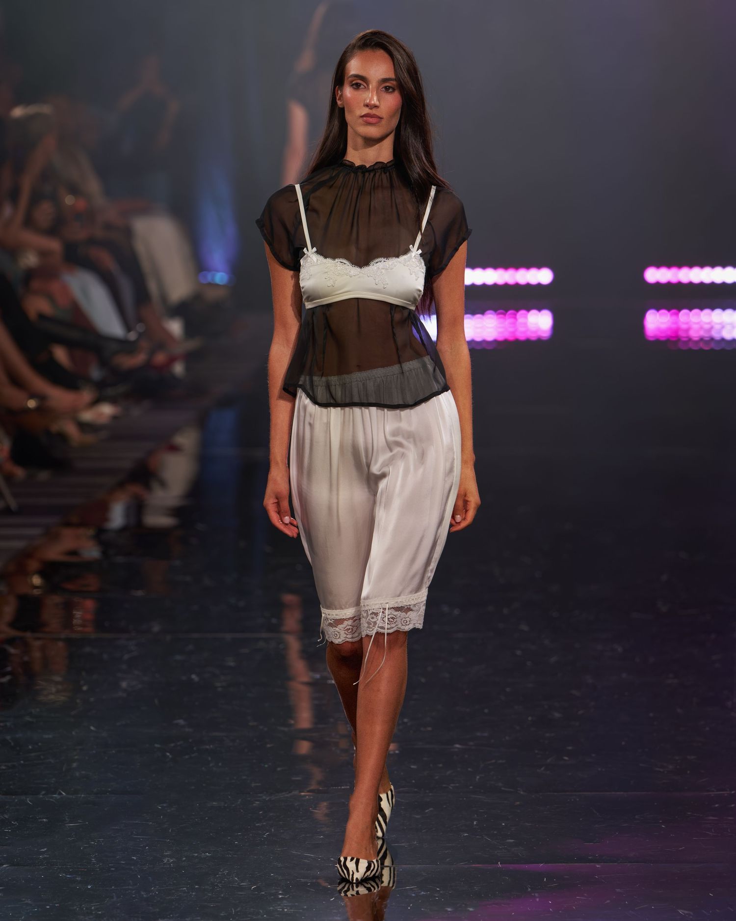 Model walks runway in sheer dark top over white lace bralette and slip shorts at a fashion show.