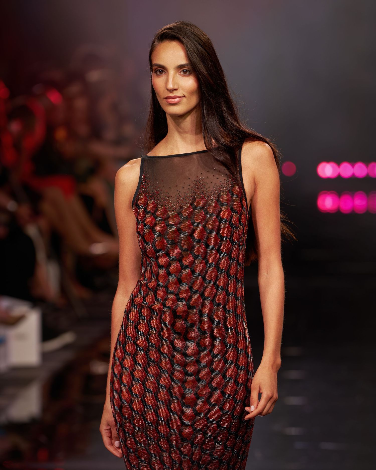 A model walks the runway in a sleeveless red and black patterned dress with sheer neckline detail.