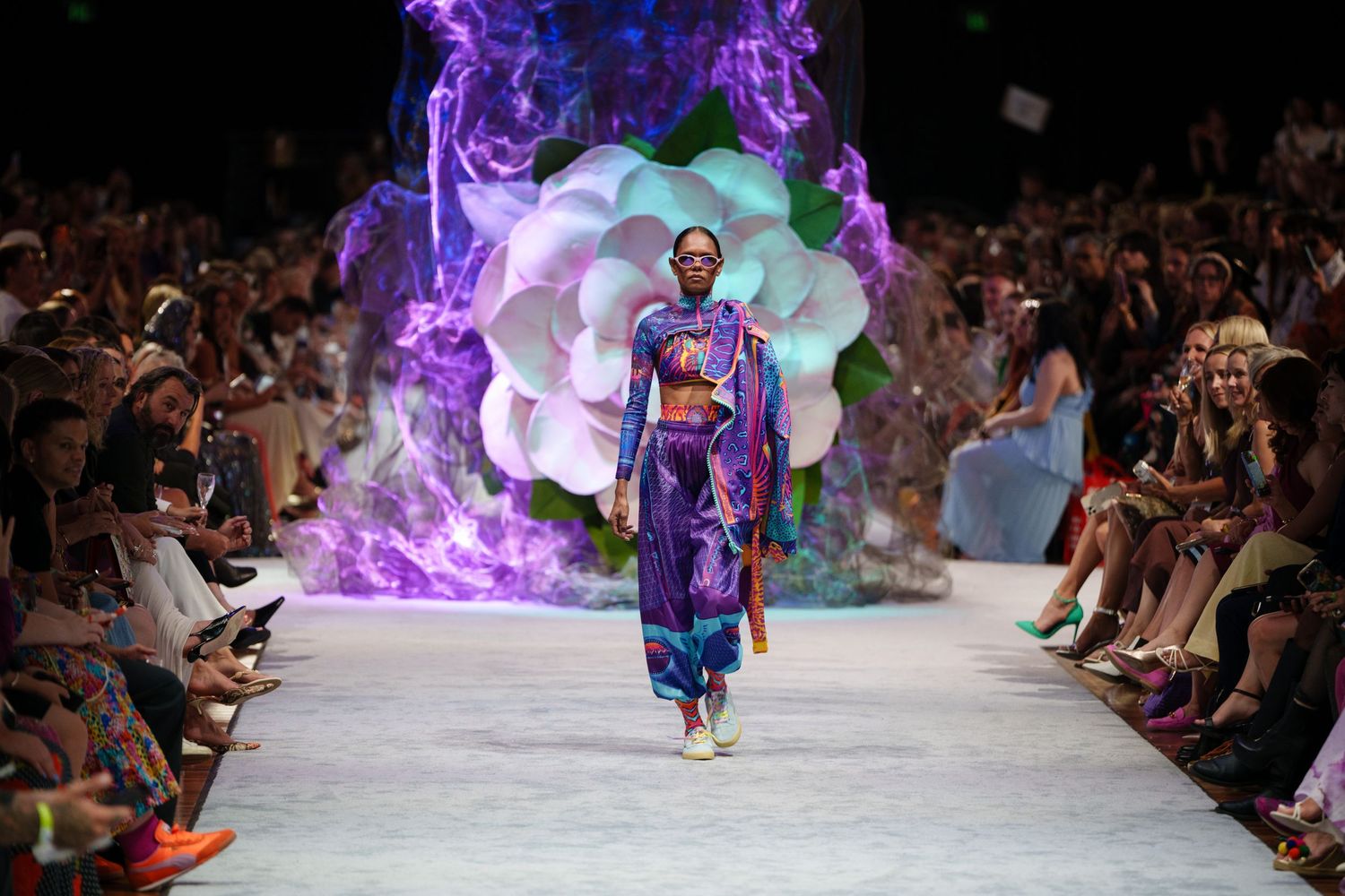 Fashion model walks runway in colorful kimono-inspired dress with dramatic purple smoke and floral backdrop at fashion show.