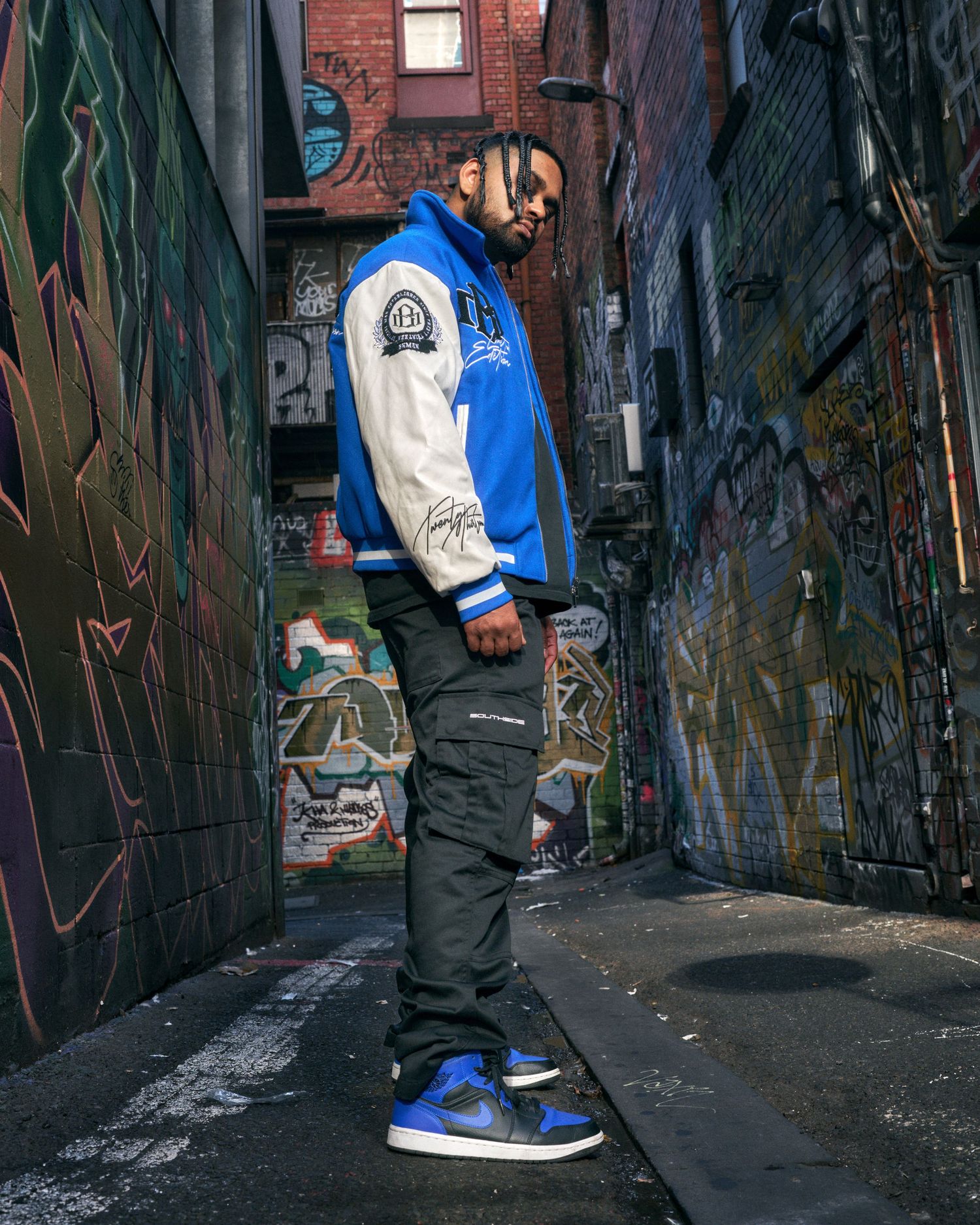 Man in blue varsity jacket and Air Jordan 1s poses in a graffiti-covered urban alley.