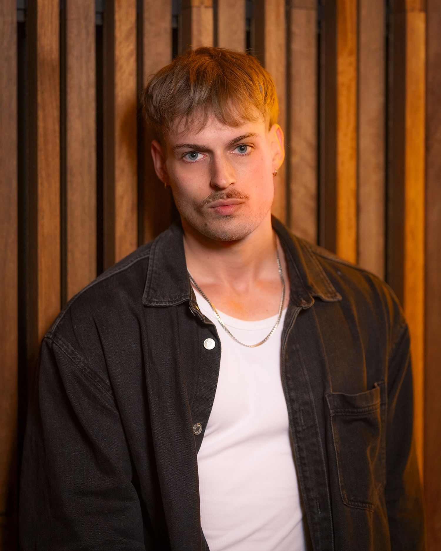 Portrait against wooden slat wall background with moody lighting and dark denim jacket over white t-shirt.