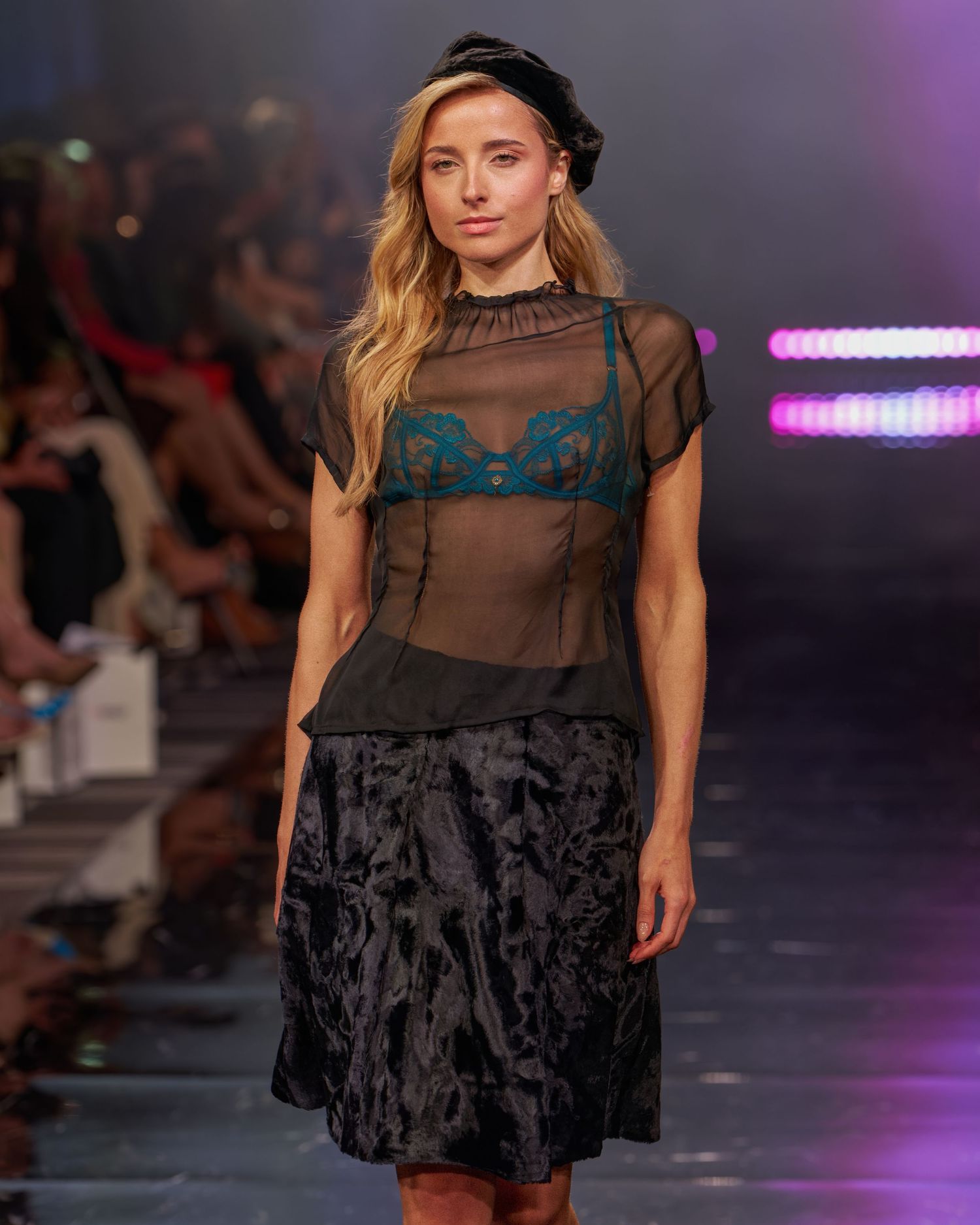 A blonde model walks the runway wearing a sheer black top over teal lace bra, black skirt, and beret hat.