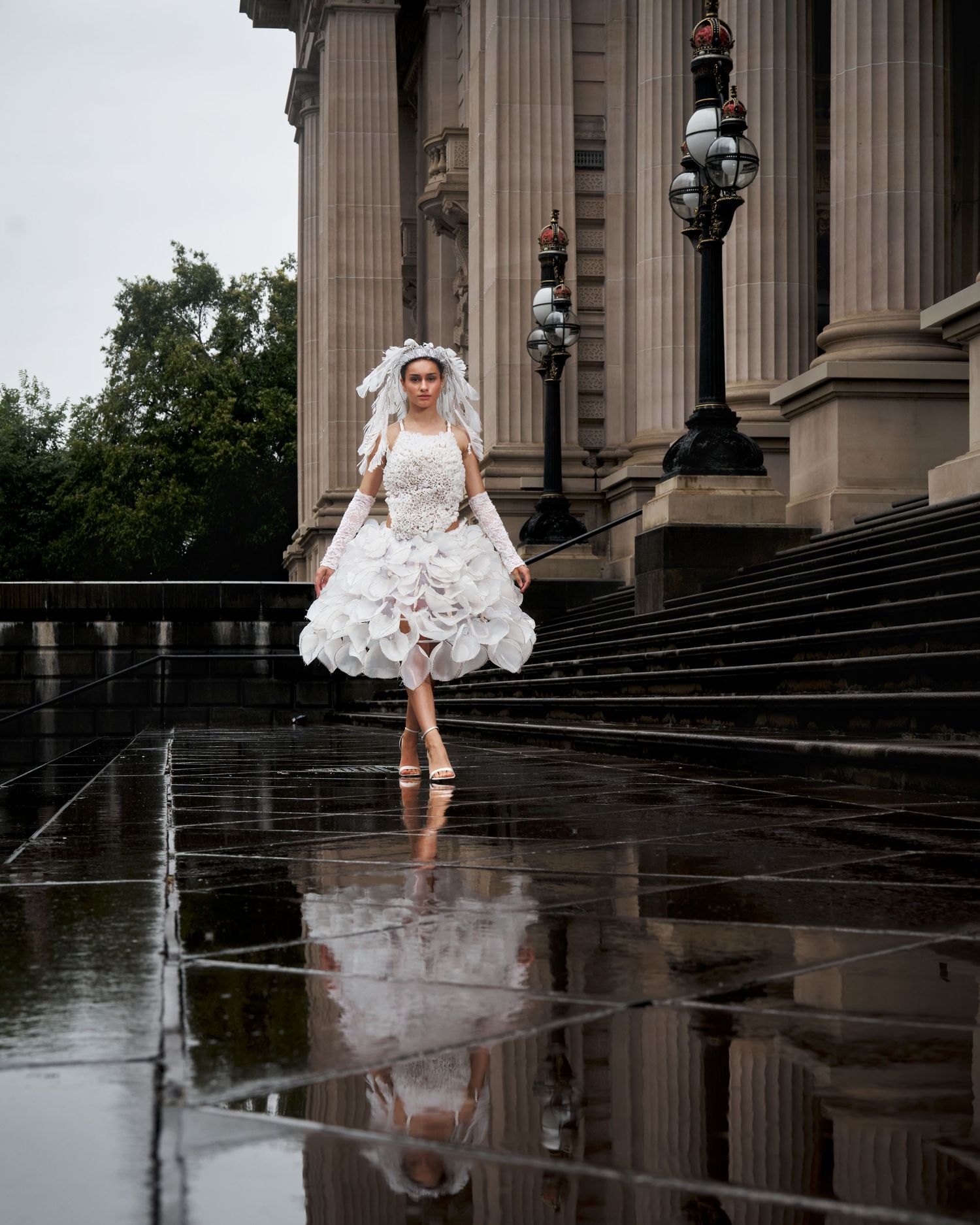 A bride in a white wedding dress playfully walks down wet stone steps reflecting in puddles near classical columns.