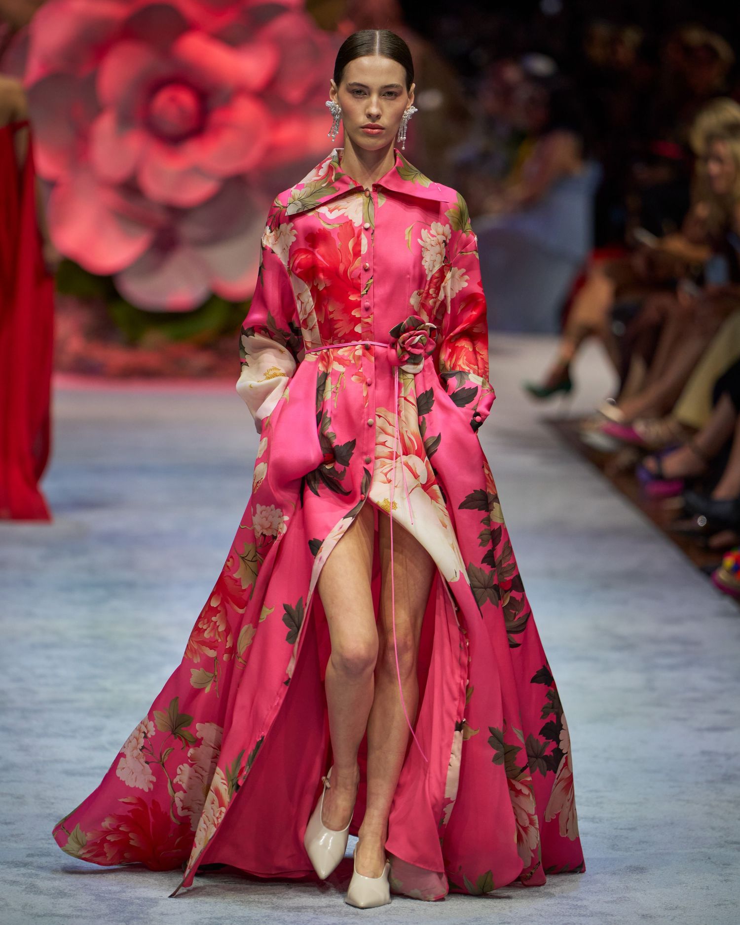 Floral pink silk gown with dramatic slit and flowing silhouette walks runway against rose-colored backdrop.