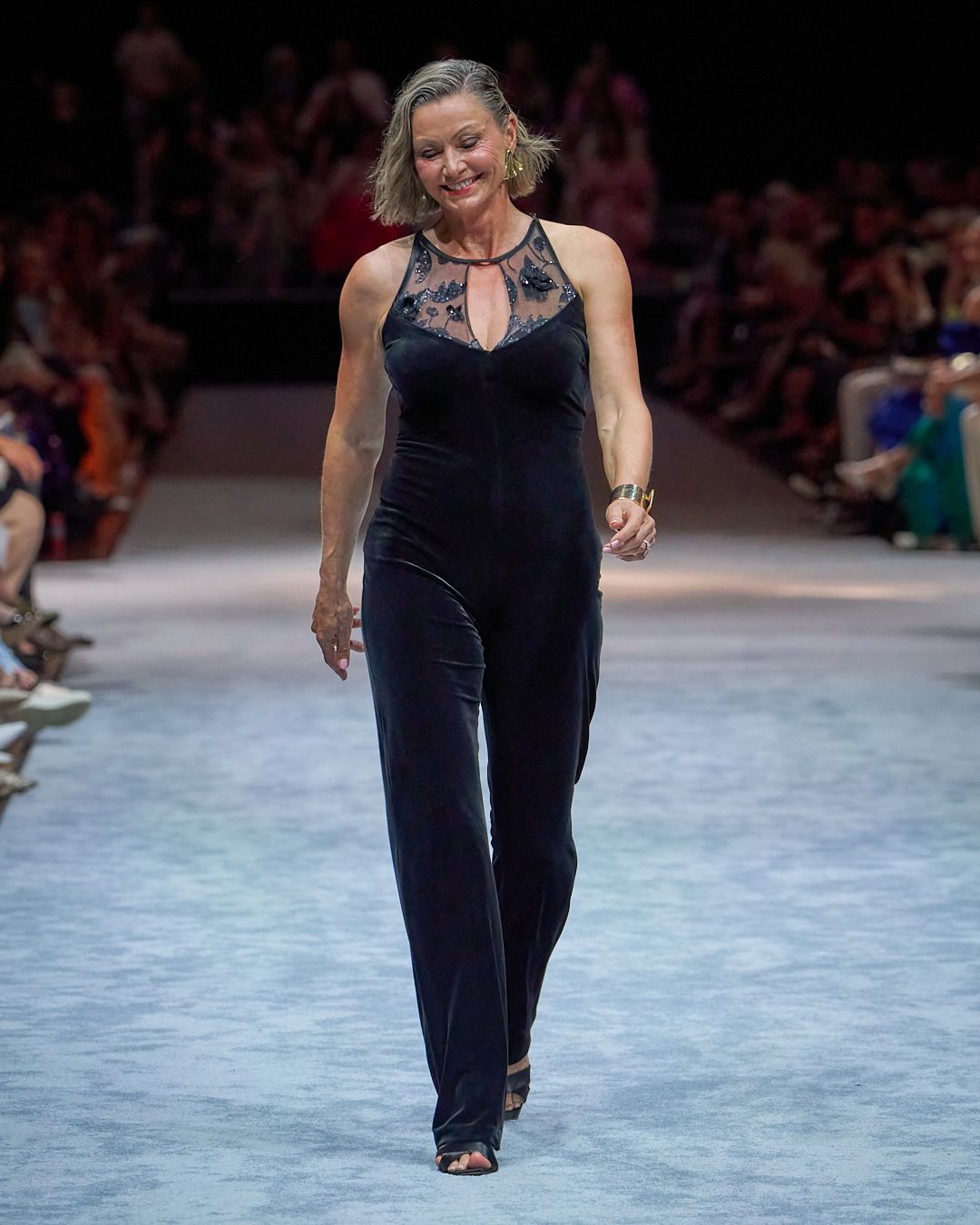 A model walks down a runway wearing a navy velvet jumpsuit with lace details during a fashion show.