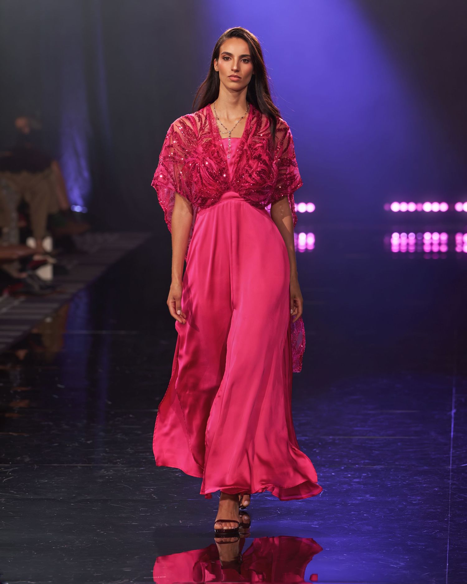 Model walks runway in a flowing hot pink maxi dress with embellished lace bodice and wide sleeves under blue stage lights.