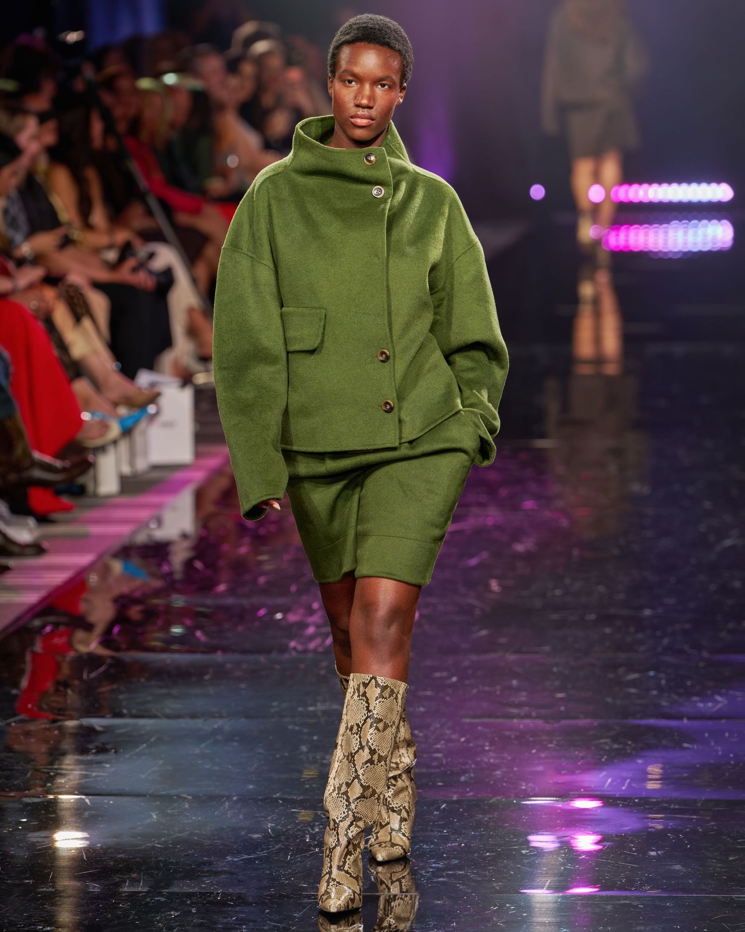 Model walks runway in olive green wool coat and matching shorts with snakeskin ankle boots at fashion show.
