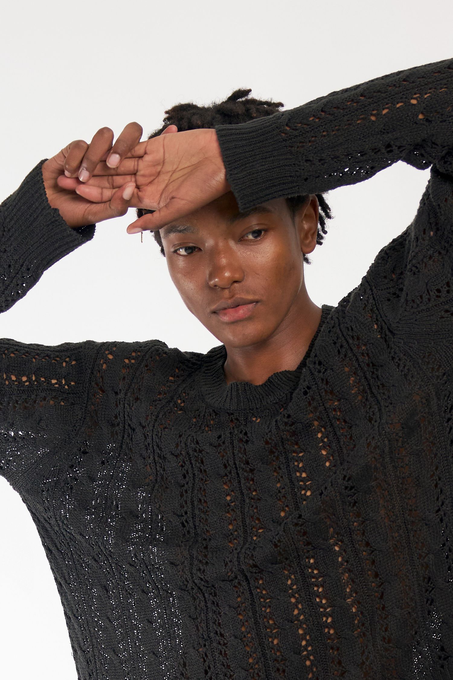 Person wearing a black openwork knit sweater poses with hands raised against white background.