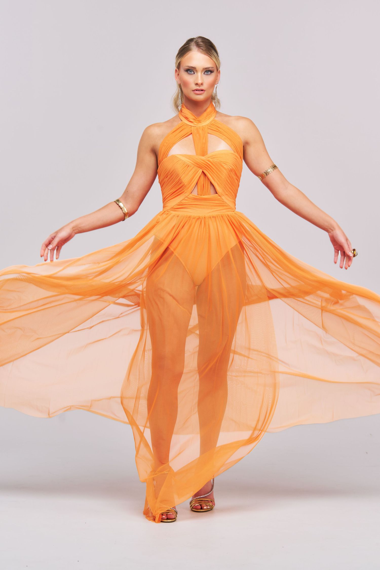 A fashion model poses in a flowing orange sheer gown with cutout details and high neckline against a gray background.