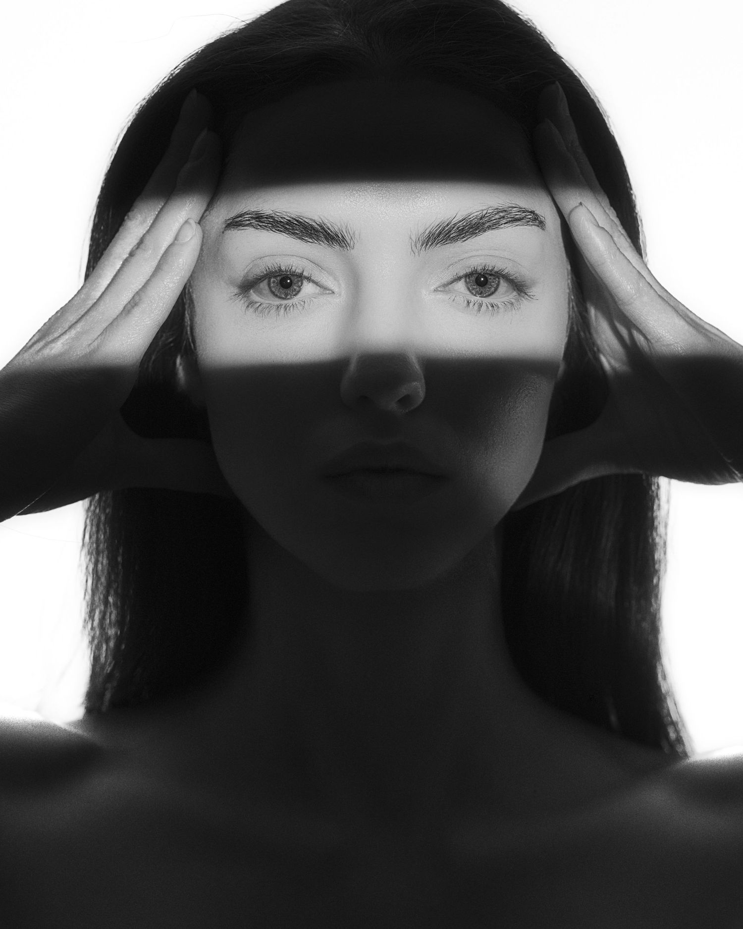 Black and white portrait of a young woman with hands on temples, dramatic light strip across her eyes.