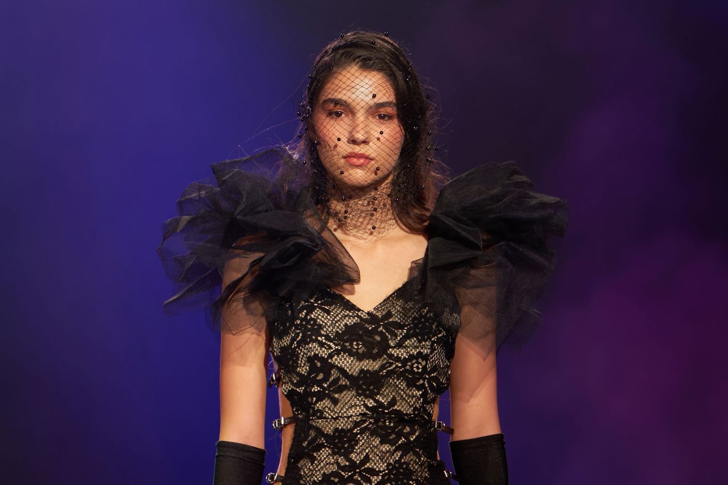 Model wearing black lace gown with tulle ruffled shoulders, veil headpiece, and gloves on fashion runway.