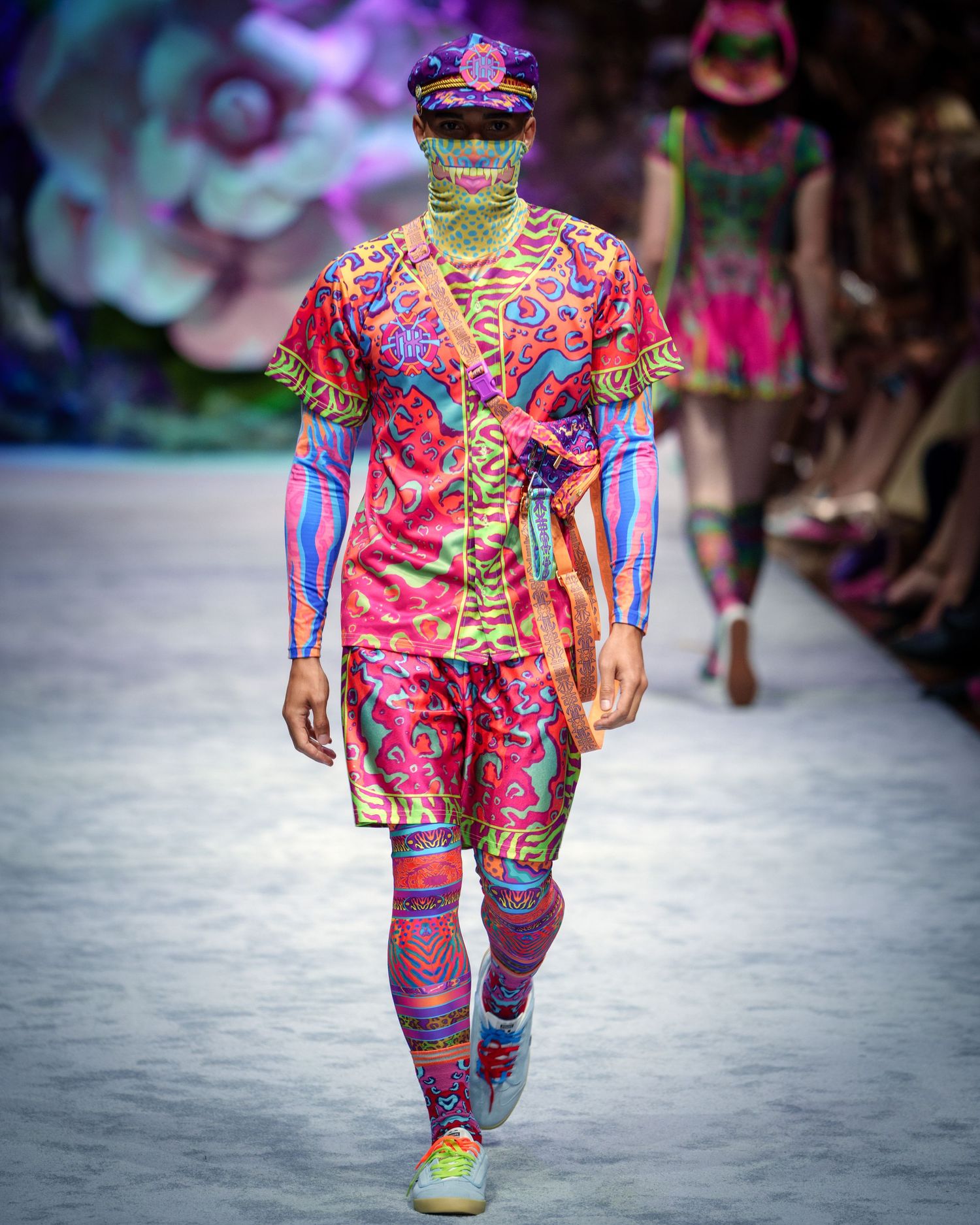 Colorful avant-garde fashion runway look featuring vibrant printed clothing with pink and blue patterns and a unique headpiece.