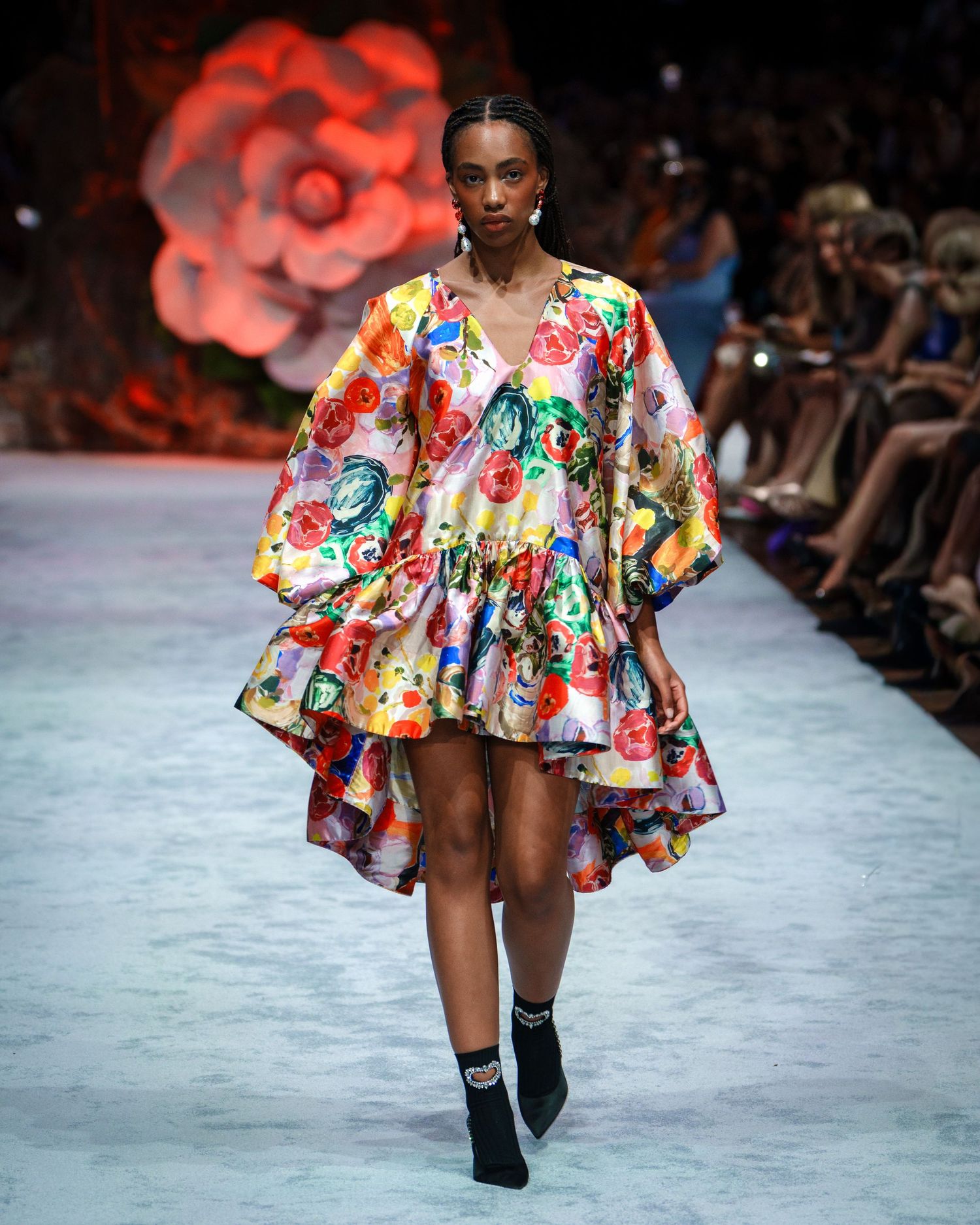 Model walks runway in colorful floral print mini dress with dramatic ruffled sleeves during fashion show.