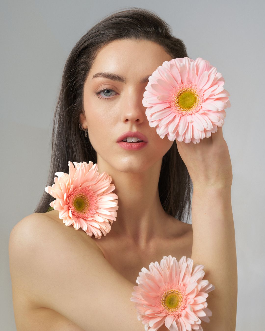 Young woman with dark hair holding three pink gerbera daisies, one covering her eye, against a gray background.