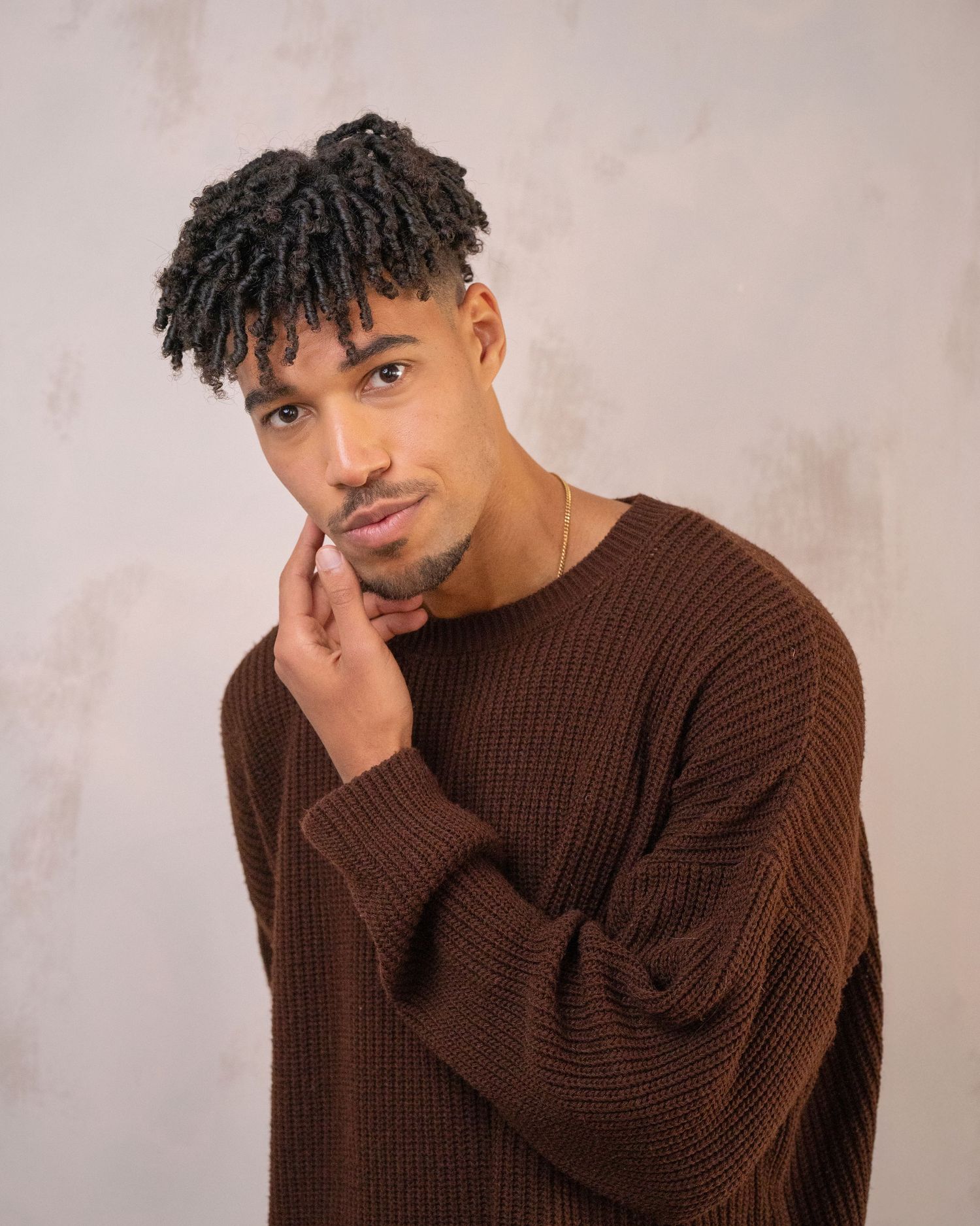 Person in a brown knit sweater poses thoughtfully against a light background.
