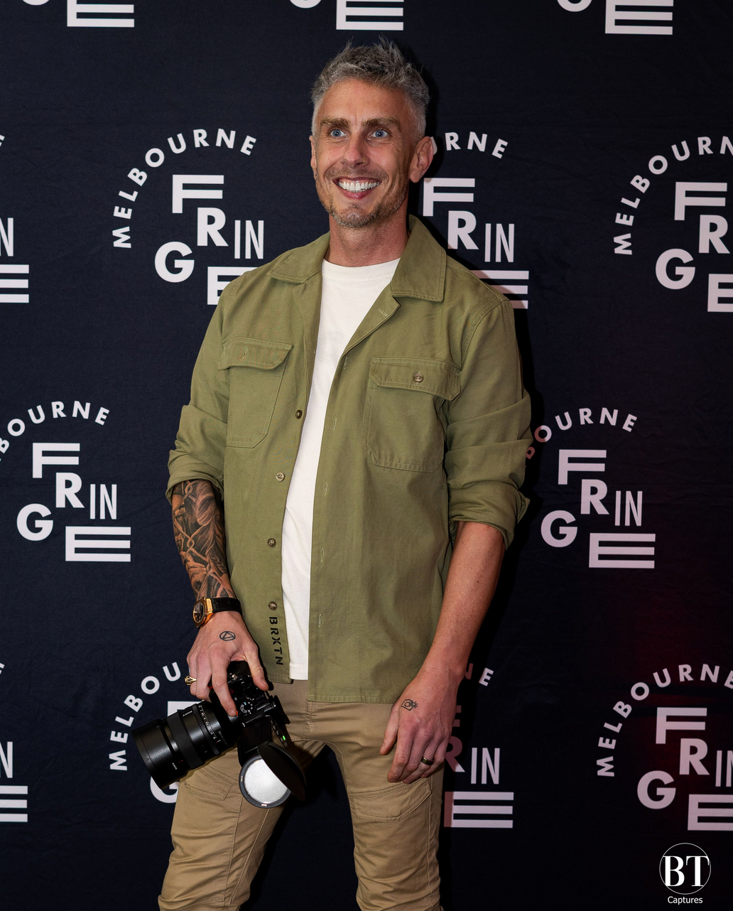 Person wearing a khaki utility jacket poses at a red carpet event with Melbourne Grin branding in the background.