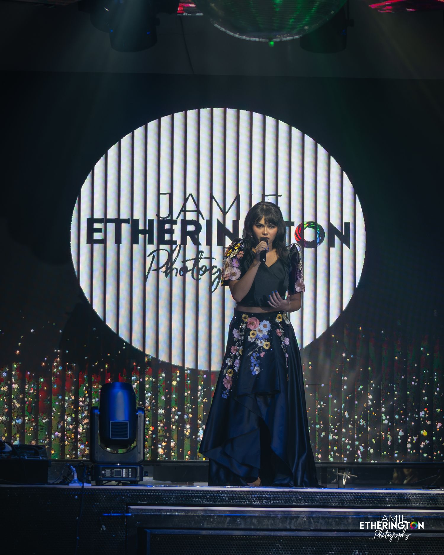 A performer sings on stage with a large illuminated circular logo and twinkling lights in the background.