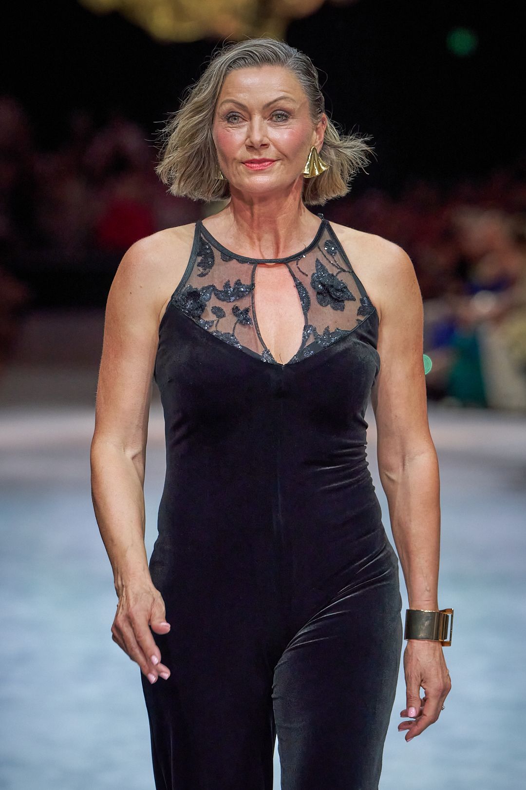 A person wearing a sleeveless black velvet dress walks confidently on a runway.