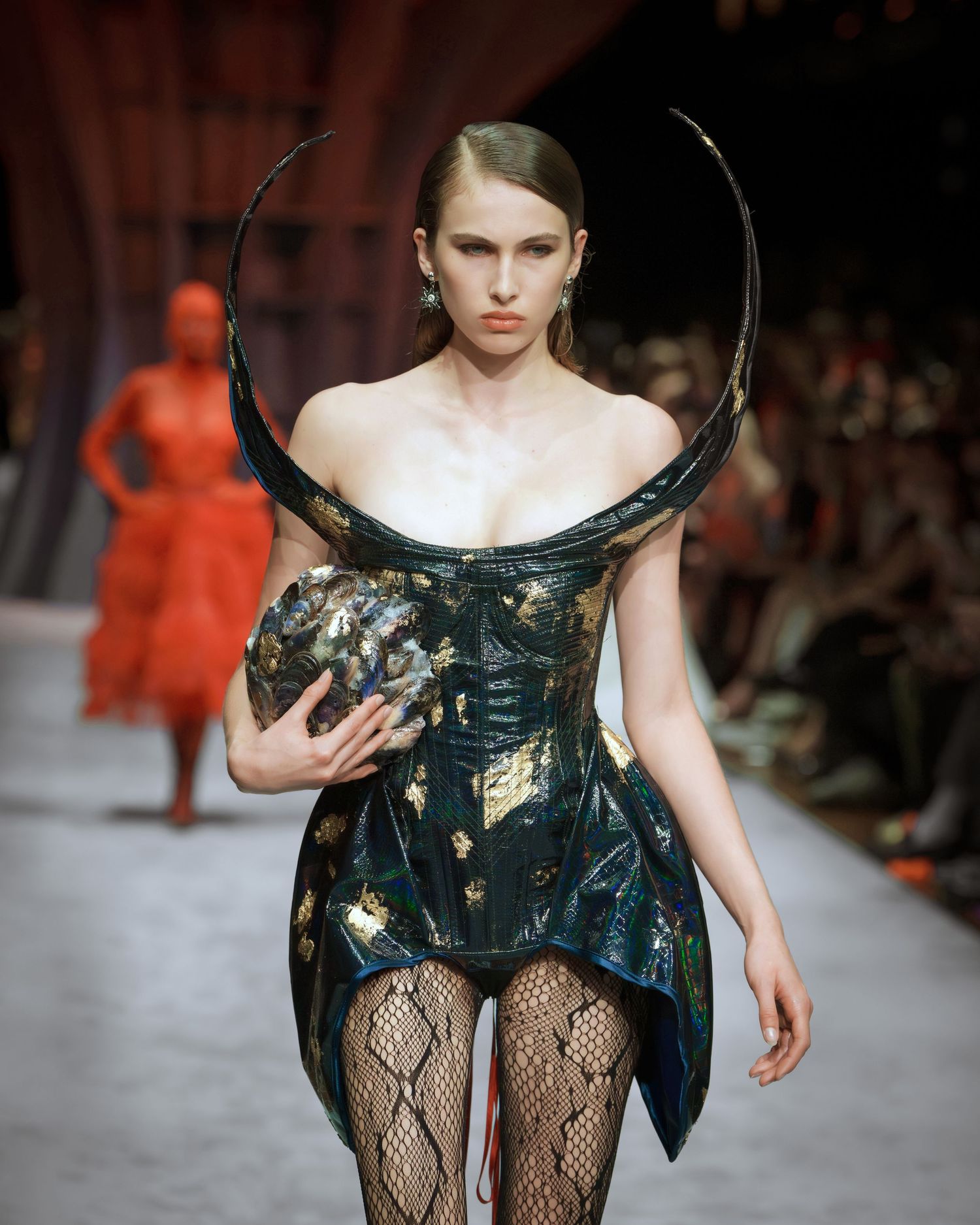 A model walks the runway in an avant-garde dress made of dark metallic material with fishnet stockings.