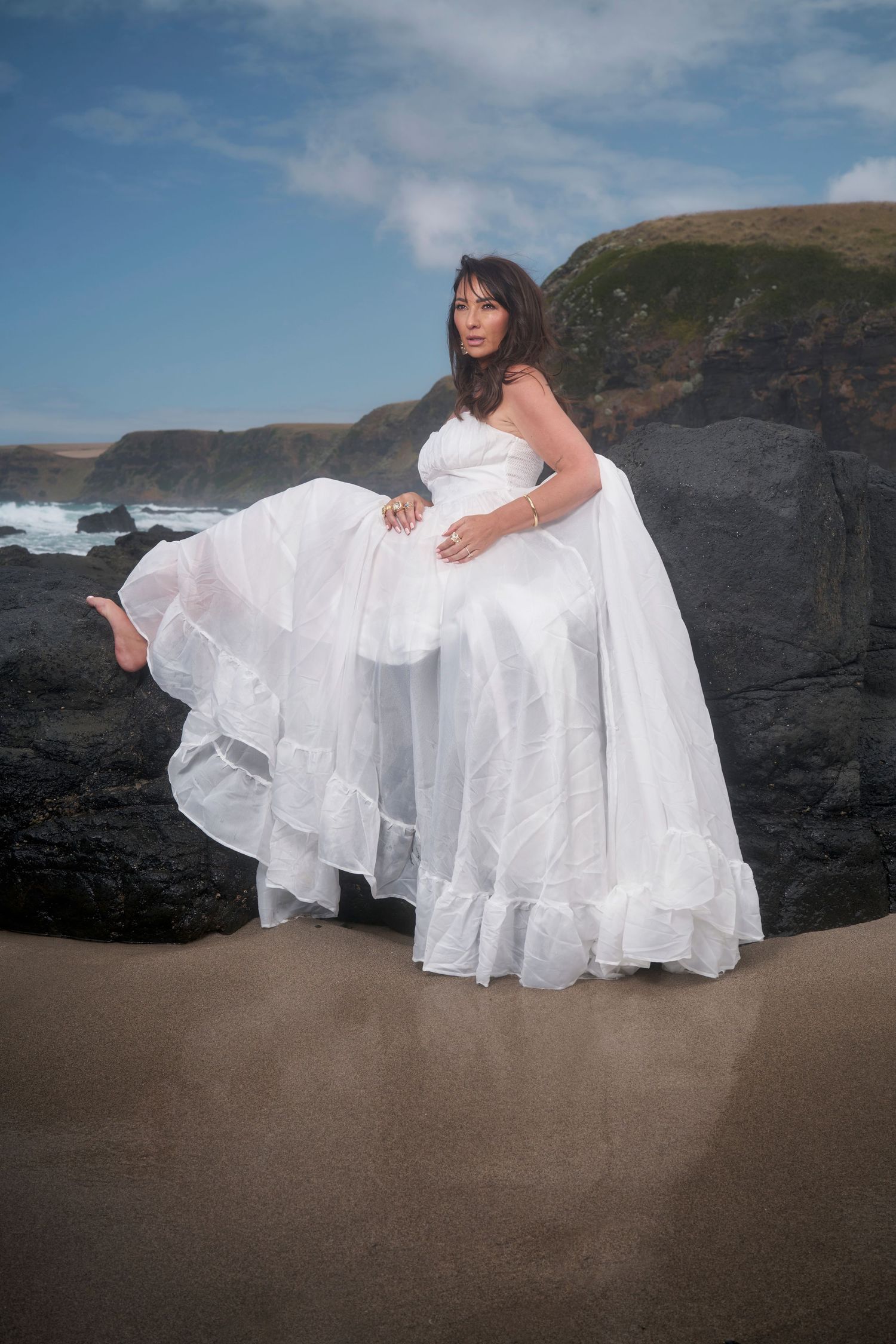 A bride in a flowing white wedding dress poses on a dramatic beach with rocky cliffs in the background.