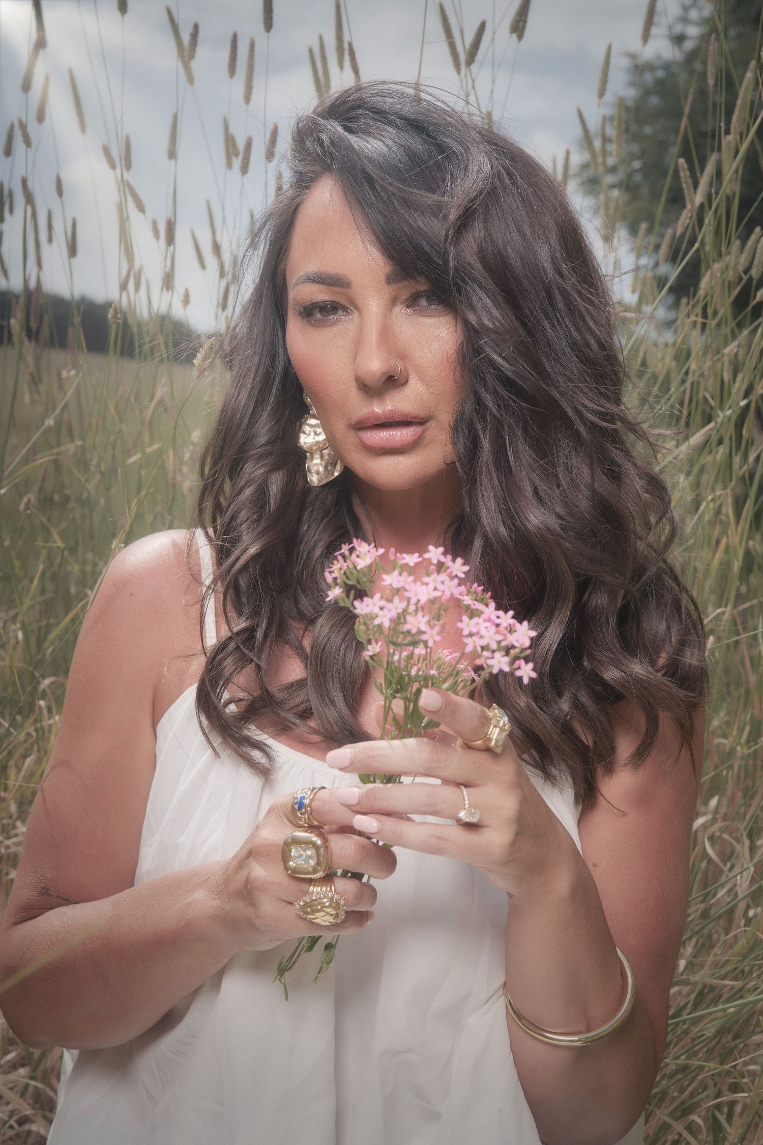 A portrait in a field of tall grass shows a person in white holding delicate pink flowers with statement jewelry.