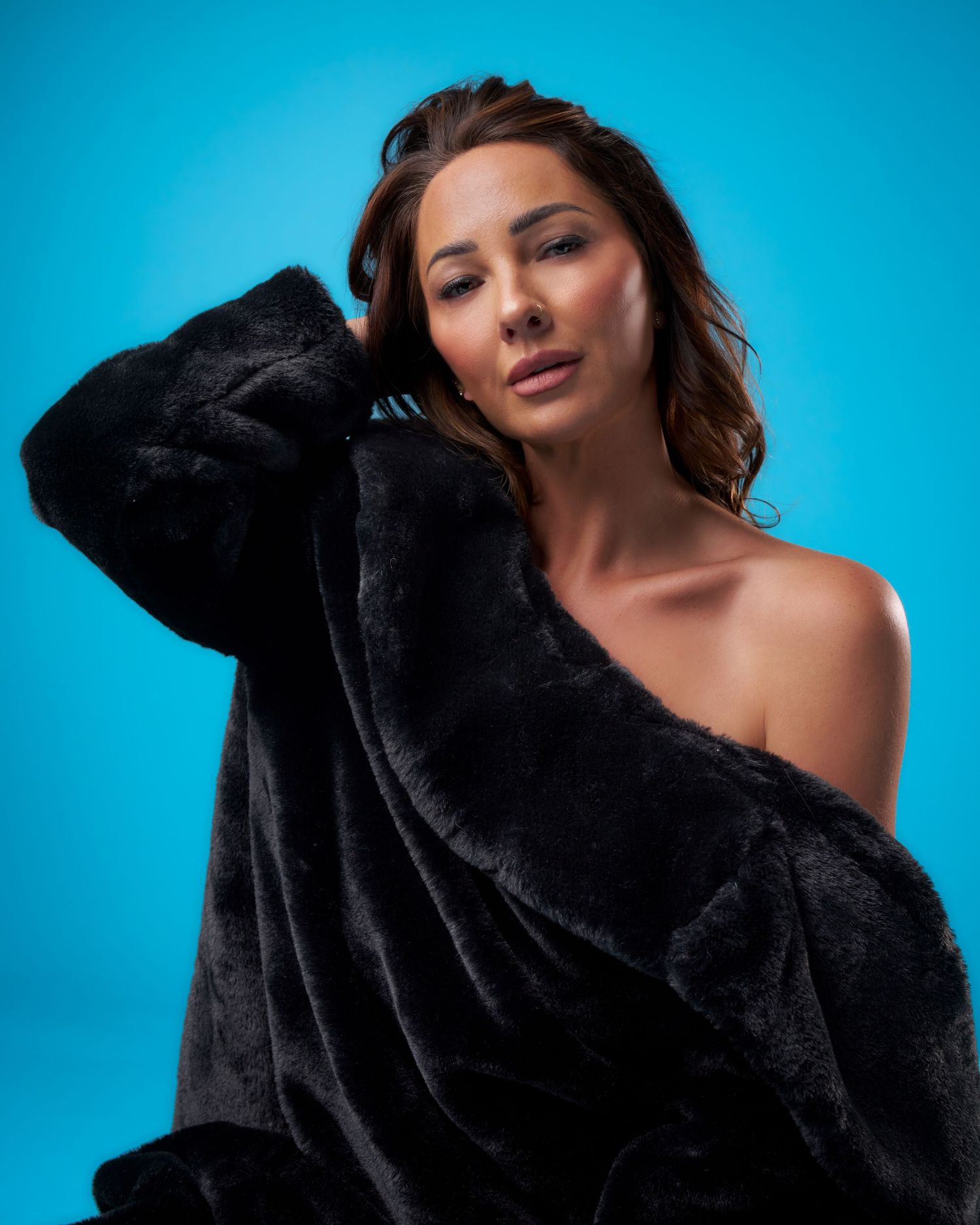 Woman in black faux fur coat posing against blue background with one shoulder exposed.