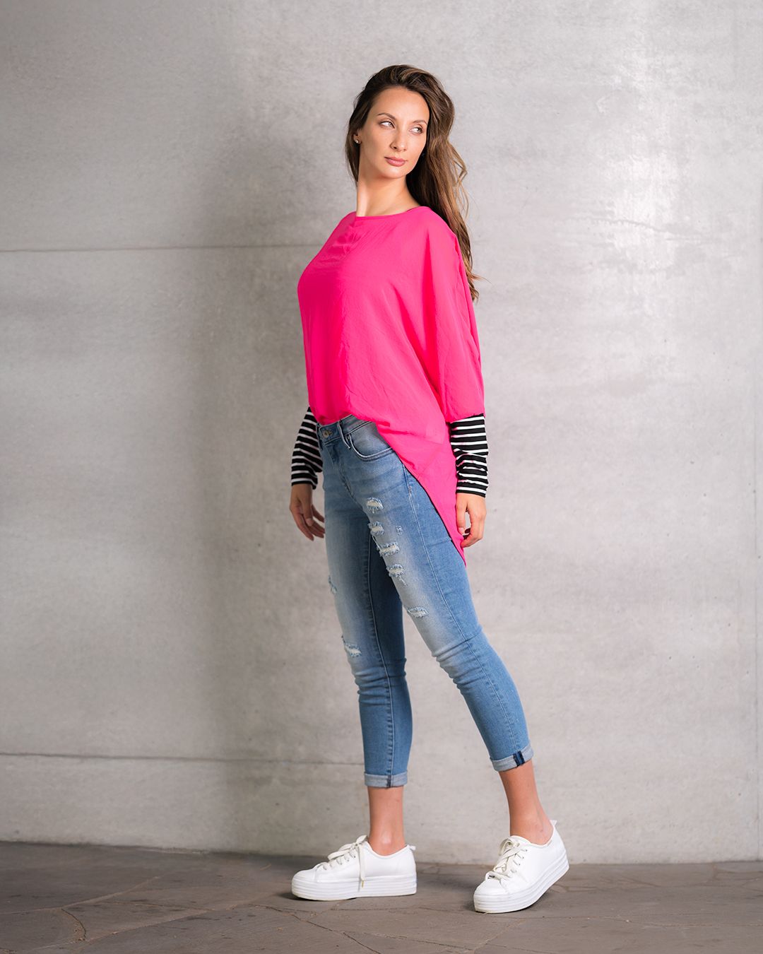 Person wearing bright pink top with striped sleeves, cropped jeans and white sneakers poses against concrete wall.