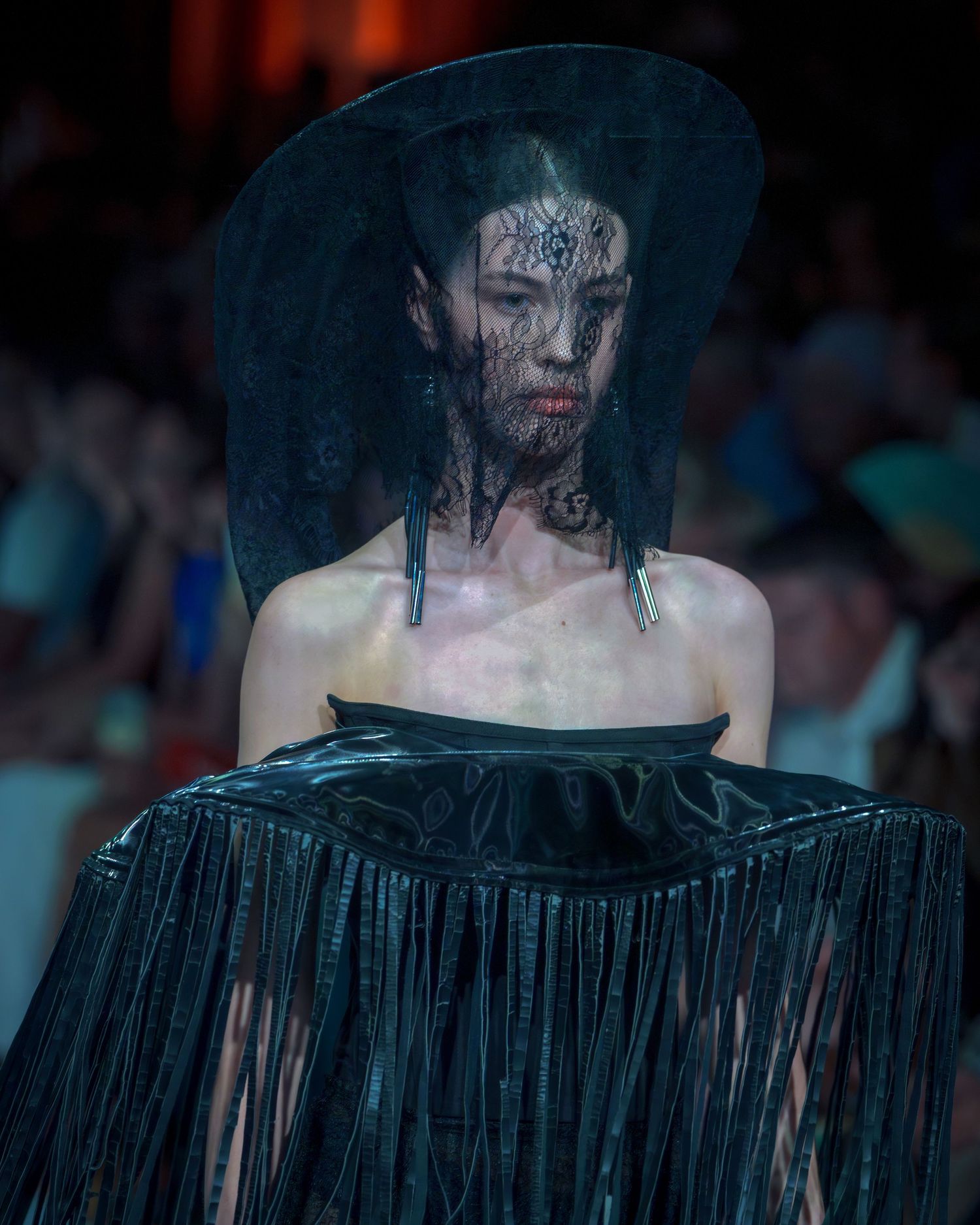 Model wearing dramatic black fringed dress and wide brim hat walks runway in dark atmospheric lighting.
