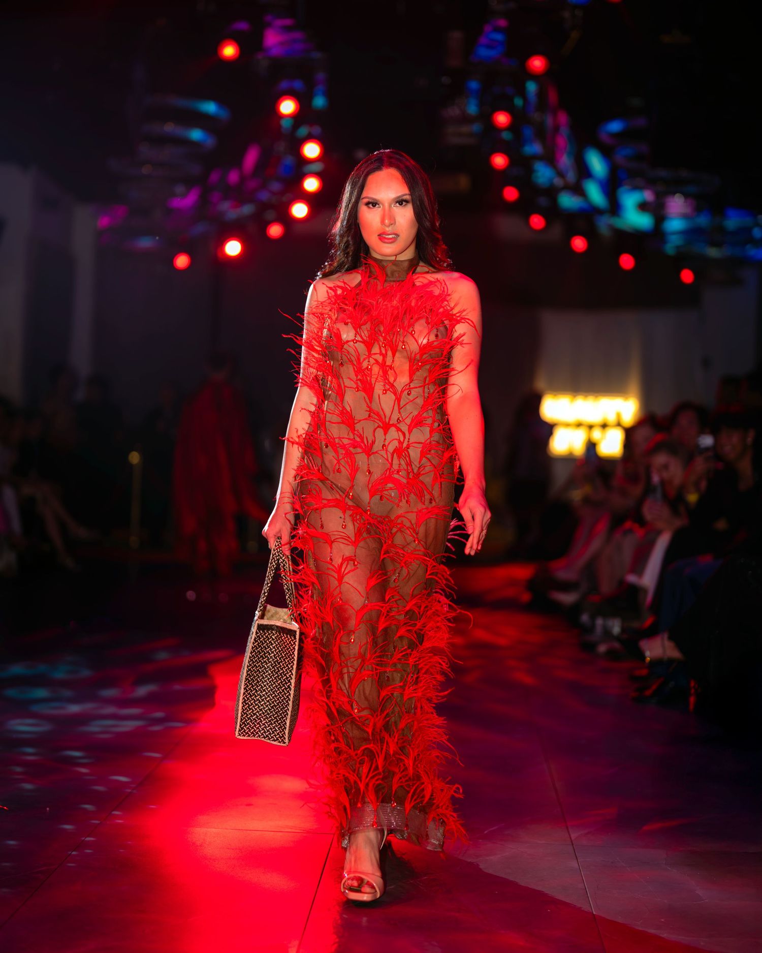 Model walks runway in flowing red feathered evening gown under dramatic lighting at fashion show.