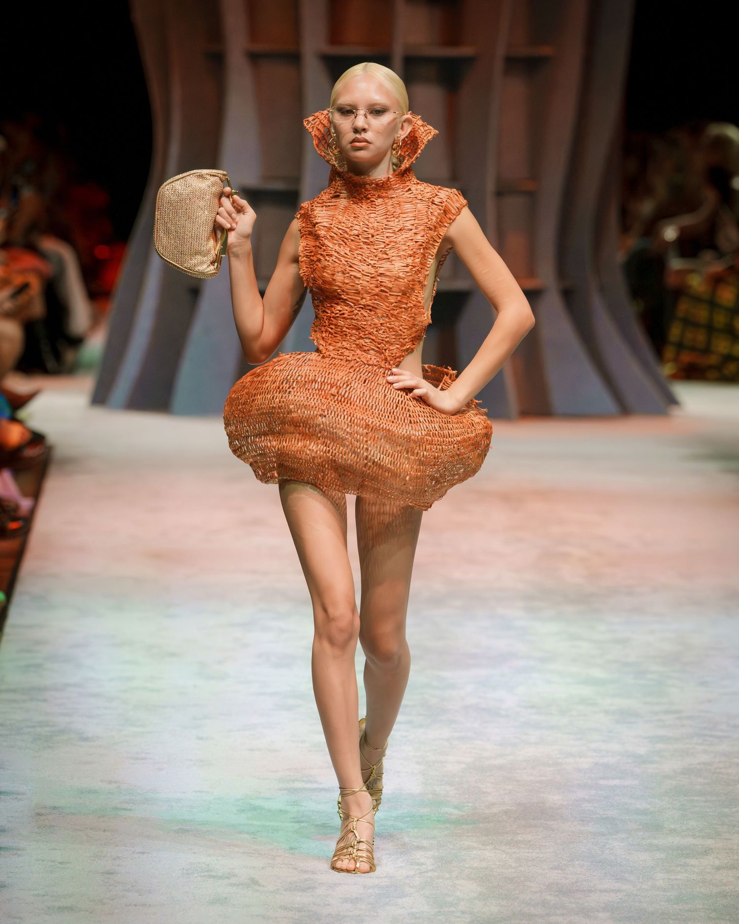 Model walks runway in orange textured mini dress and flat sandals during fashion show.
