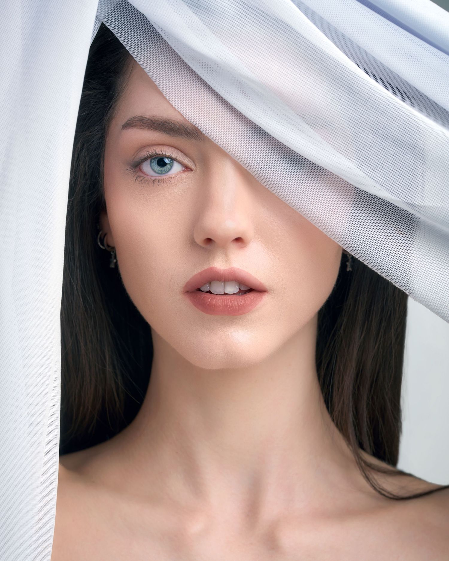 A young woman with blue eyes and dark hair partially covered by flowing white fabric in an artistic portrait.