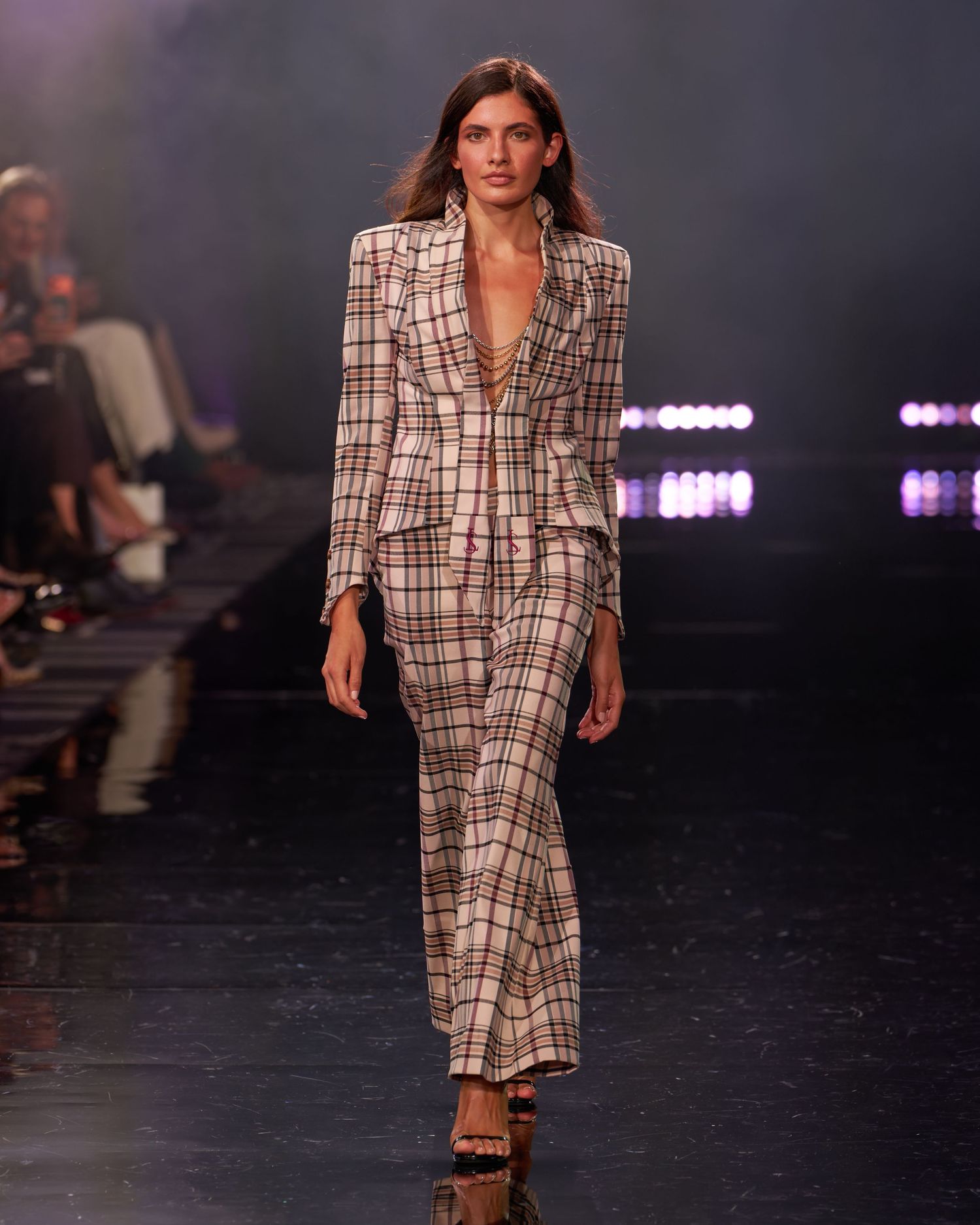 Model walks runway in plaid blazer and matching maxi skirt suit at fashion show.