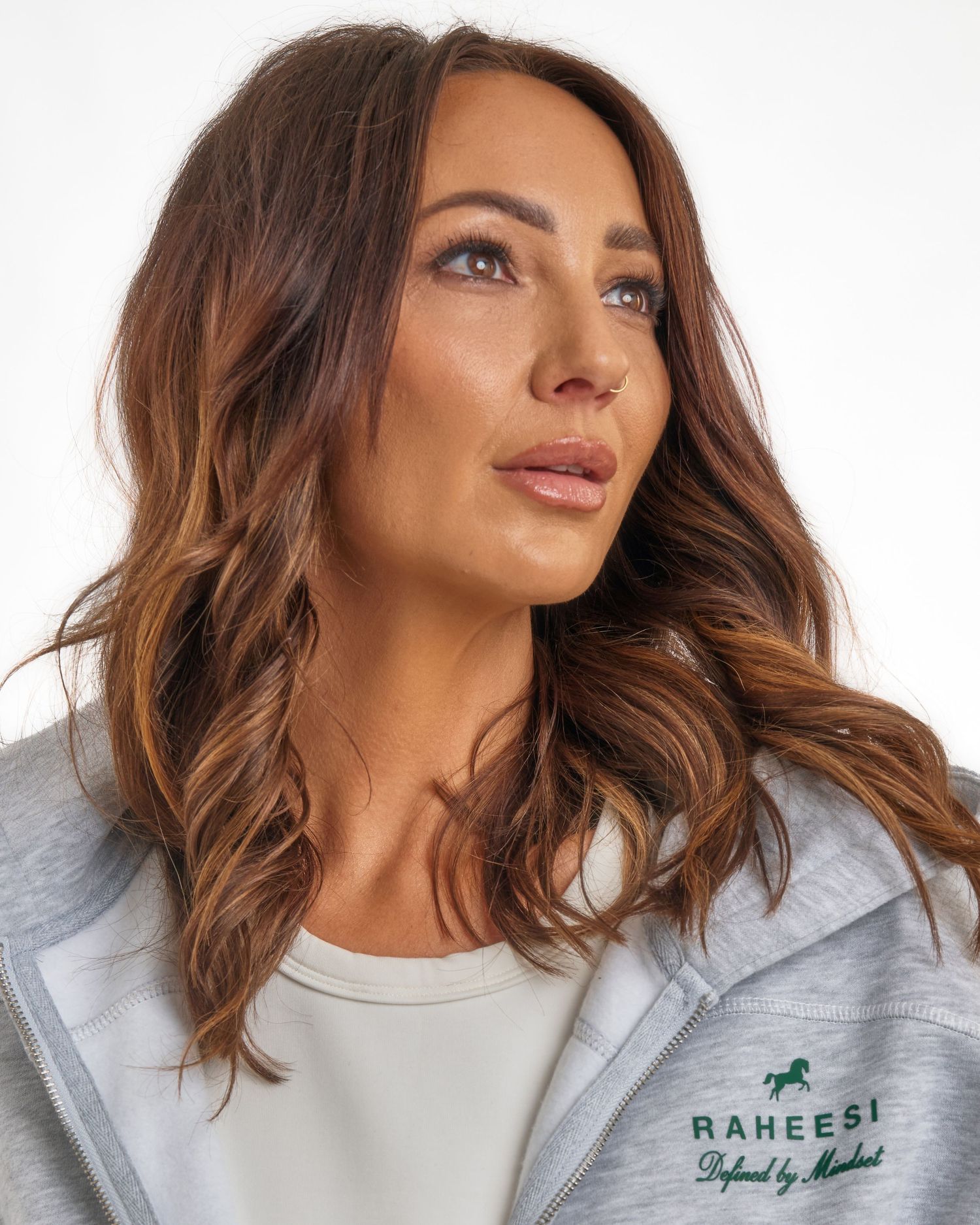 Close-up of woman with wavy brown hair wearing Raheesi branded grey hoodie looking upward.
