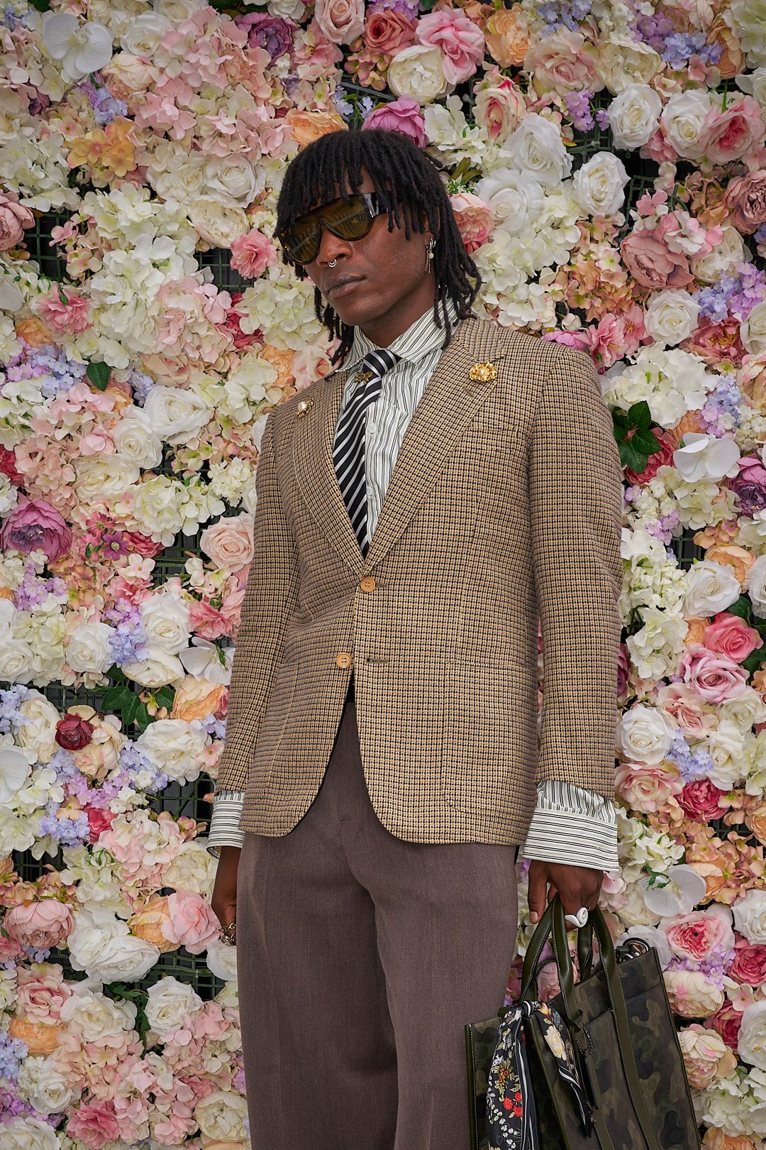 A figure in a brown tweed blazer and striped tie poses against a wall of pink and white flowers.