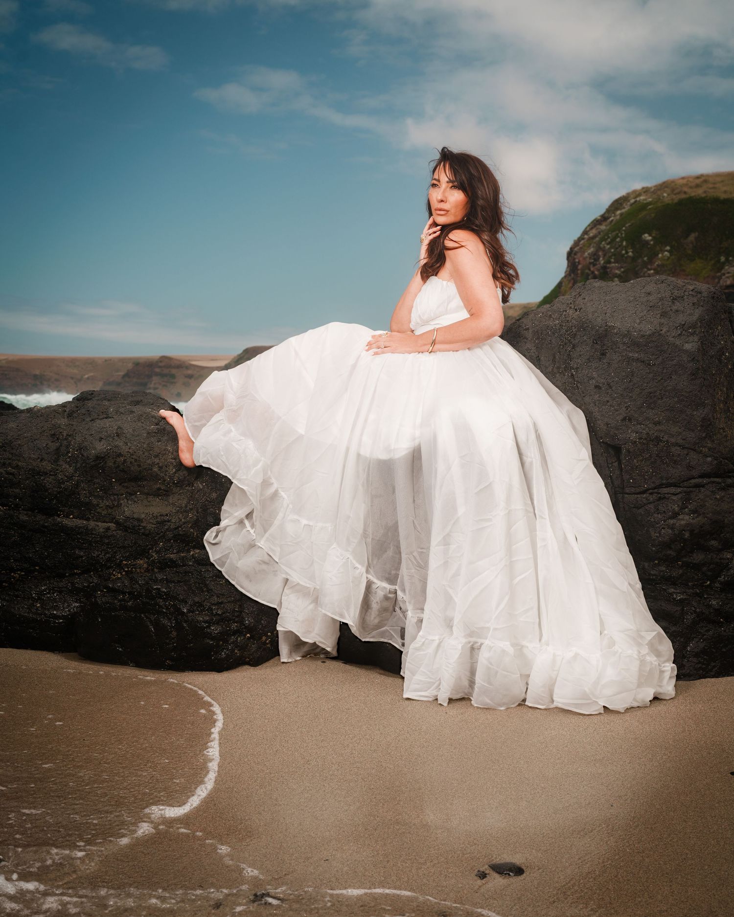 A flowing white wedding dress billows in the wind against dramatic coastal rocks and sandy beach at sunset.