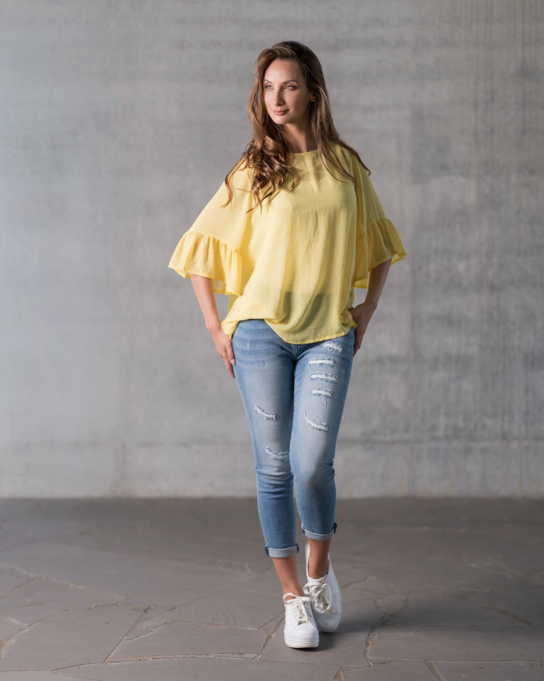 Woman in yellow flutter sleeve top and distressed jeans poses against grey wall in casual summer outfit.
