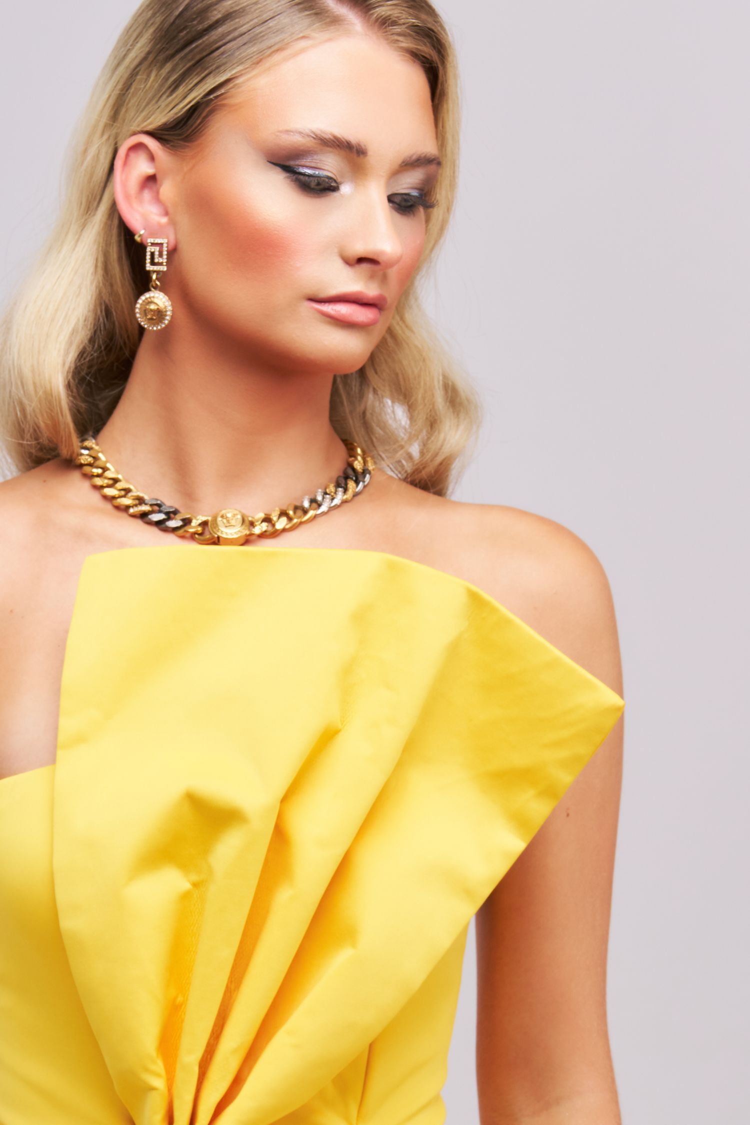 A woman in a bright yellow evening dress with ruffled details and gold chain necklace poses against a gray background.