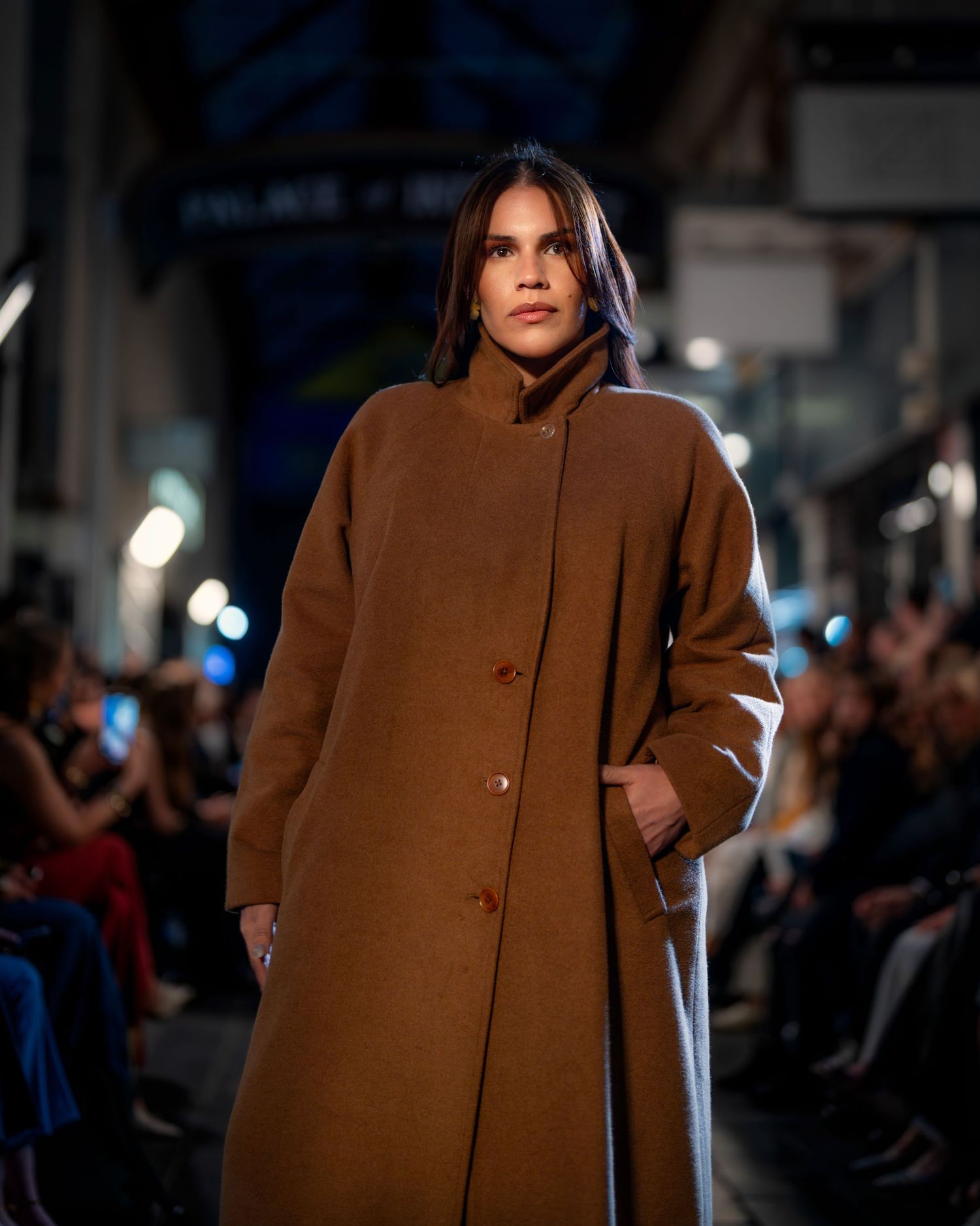 Model walks the runway wearing a brown oversized coat during an evening fashion show presentation.
