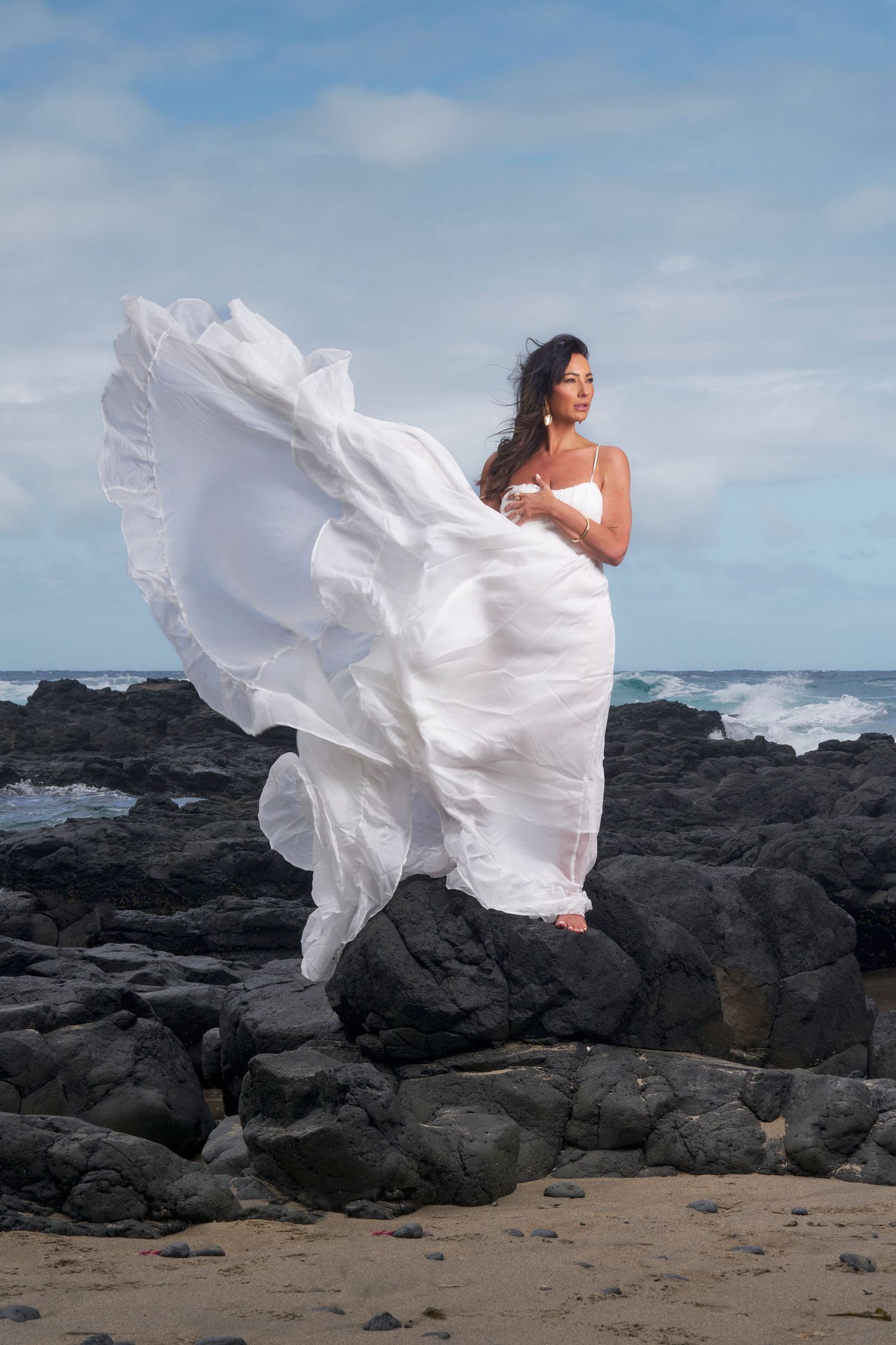 A flowing white wedding dress billows dramatically against dark volcanic rocks and ocean waves on a beach.