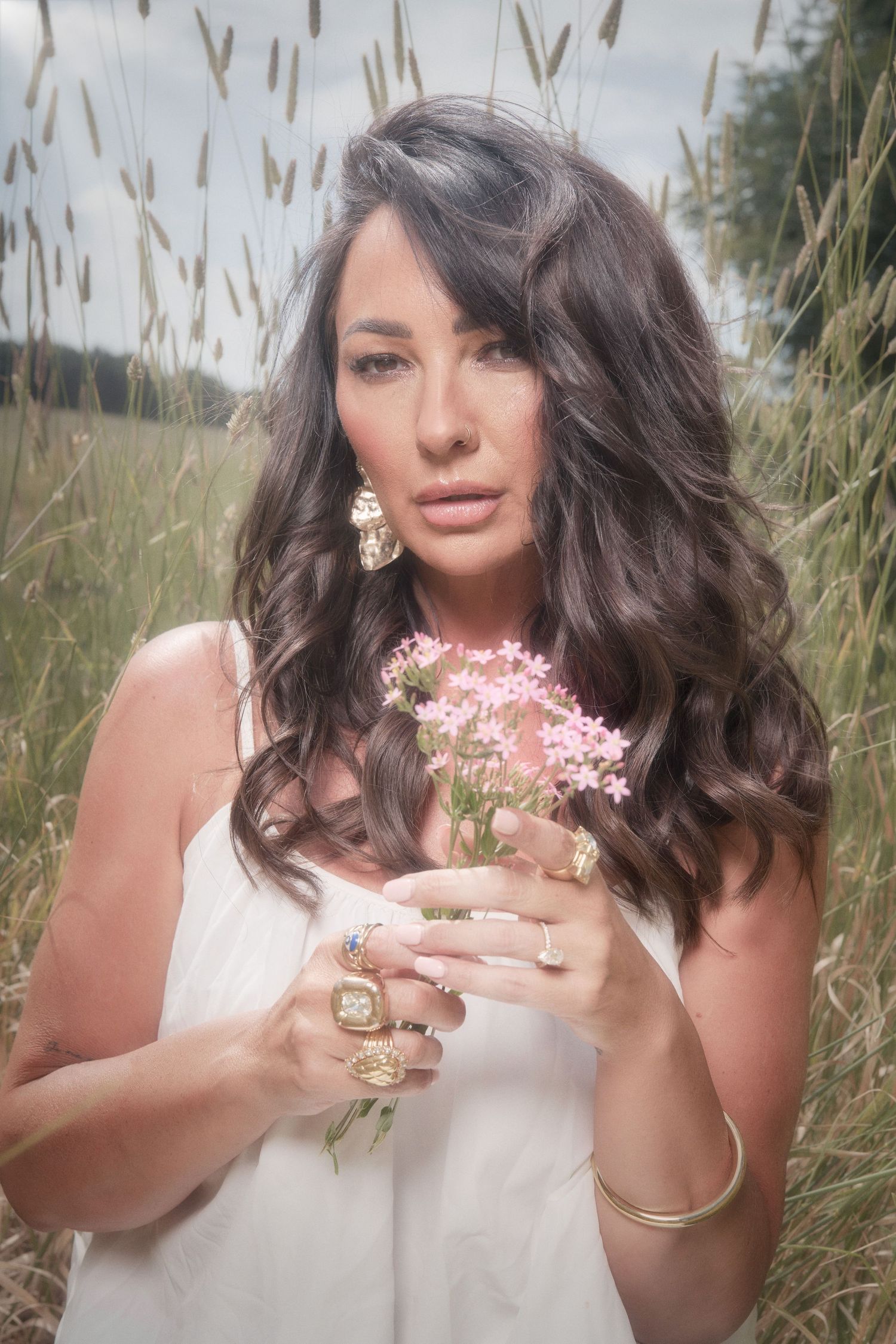 Ethereal portrait in a wheat field features long dark wavy hair, white dress, pink flowers and statement jewelry.