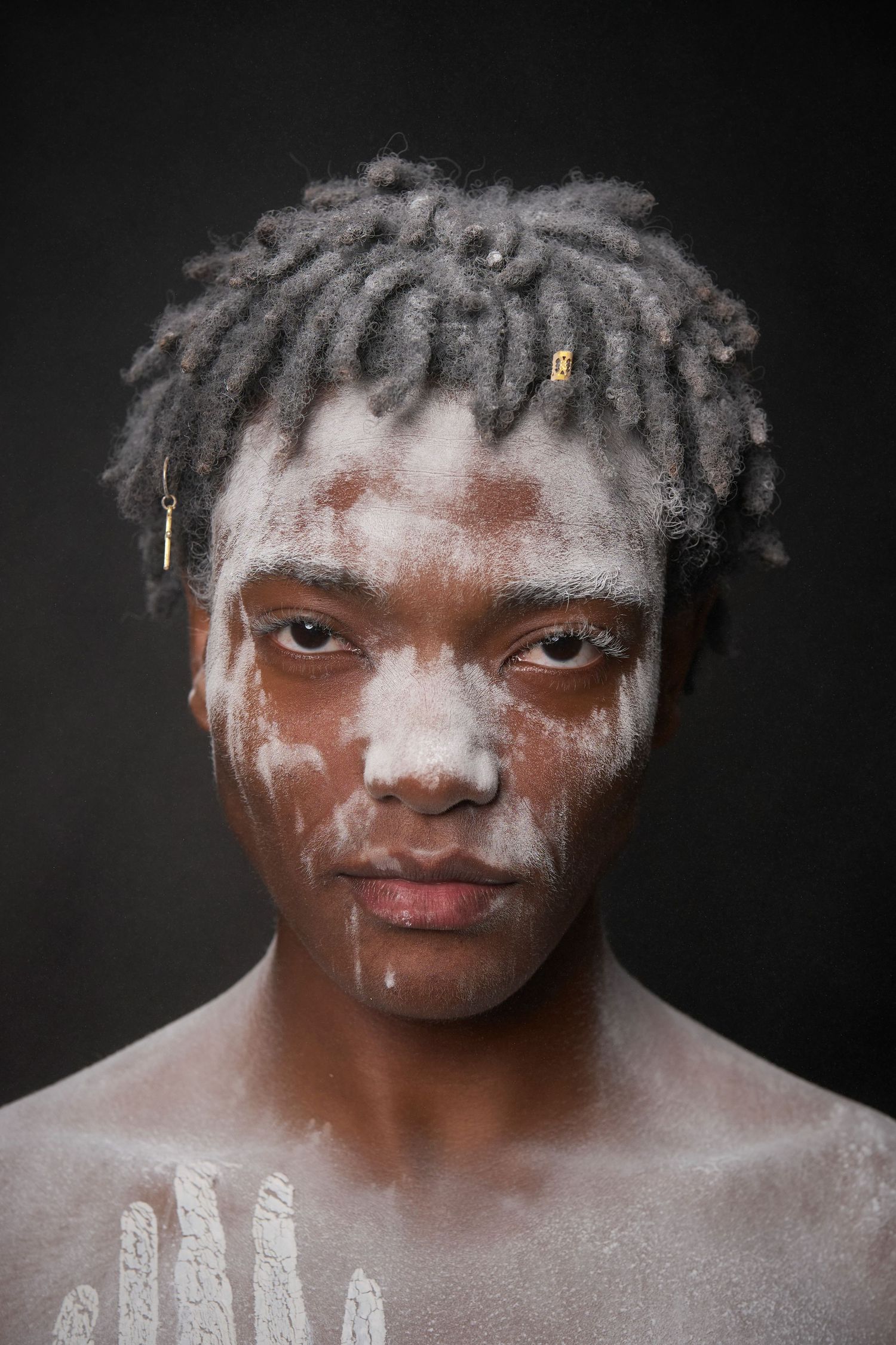 Portrait against black background showing face with white paint or powder markings and dreadlocked hair.