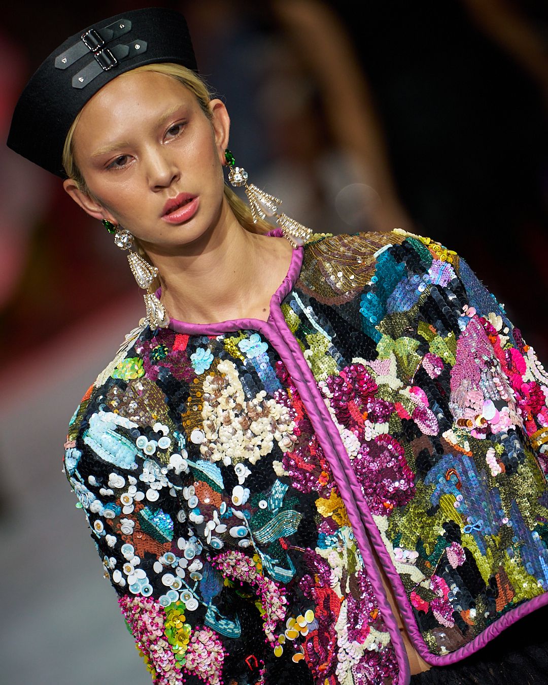 A colorful sequined fashion garment with pink, blue, and gold sparkly details shimmers on the runway.
