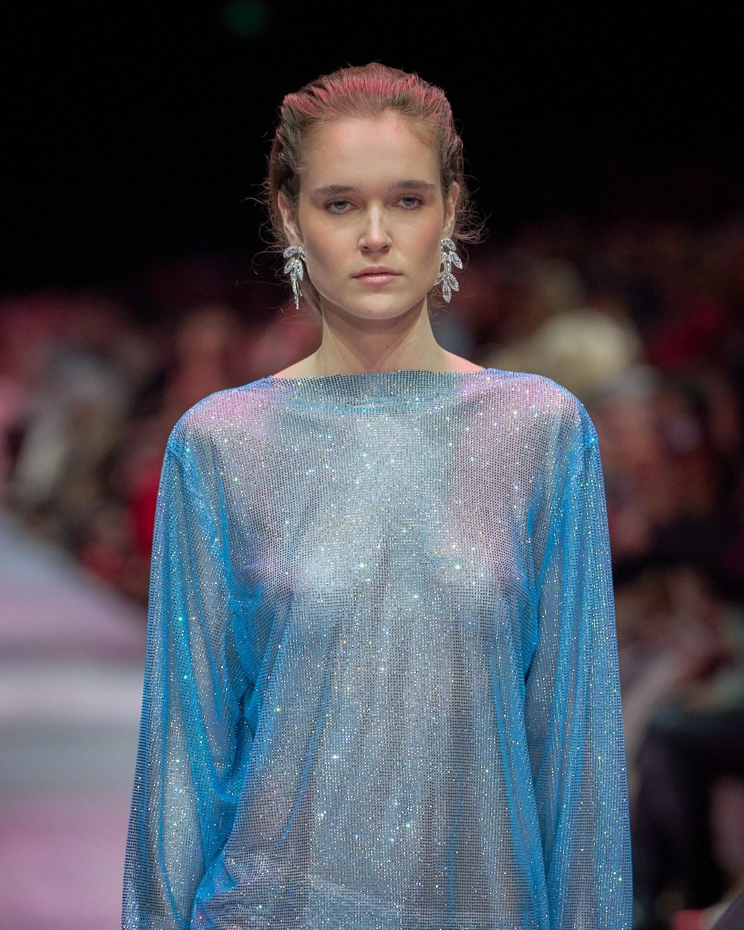 A model walks the runway wearing a sparkly blue and silver long-sleeved evening gown with a shimmering iridescent finish.