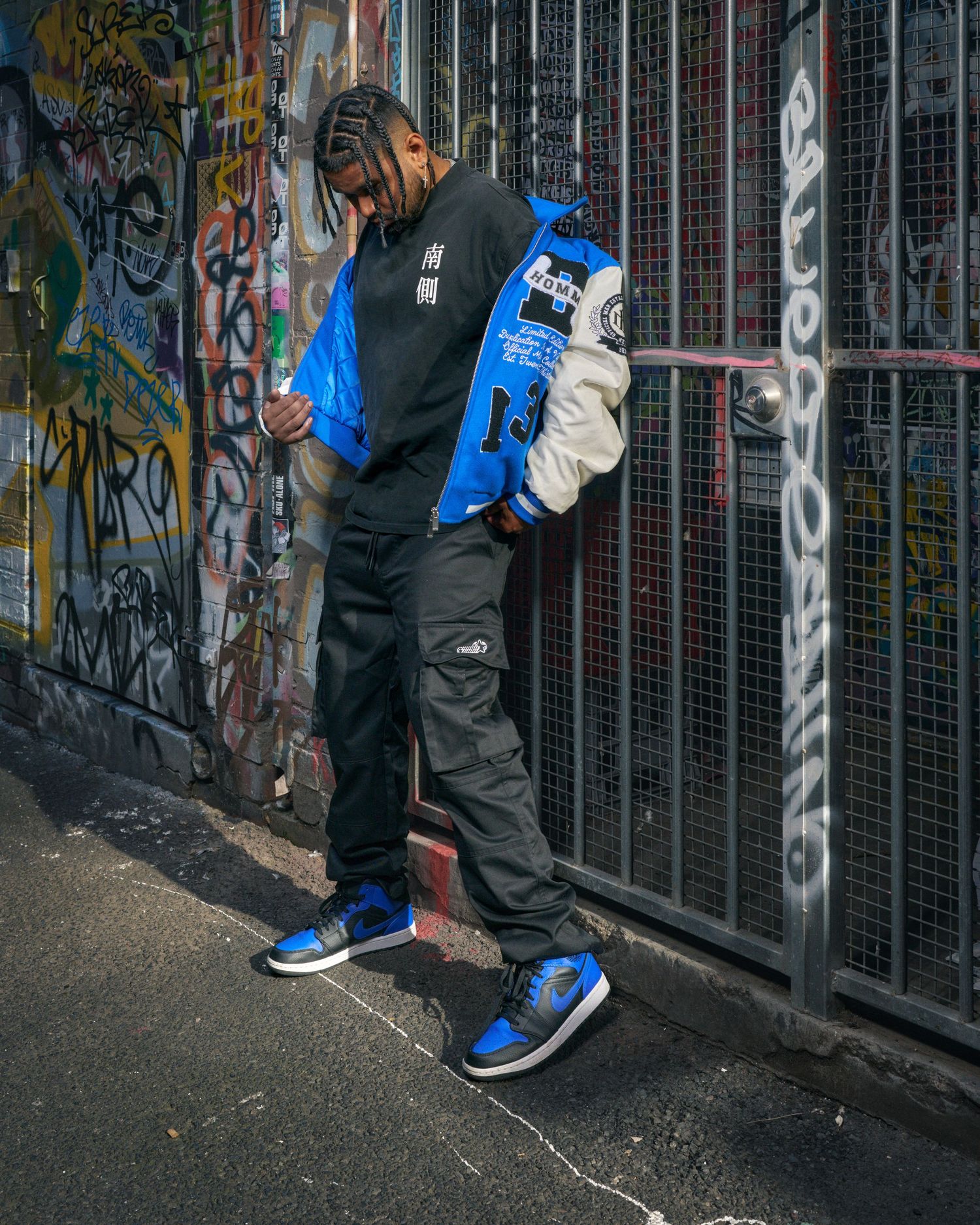 Man in blue varsity jacket, black cargo pants, and blue Jordan 1s posing against graffiti wall in urban alley.