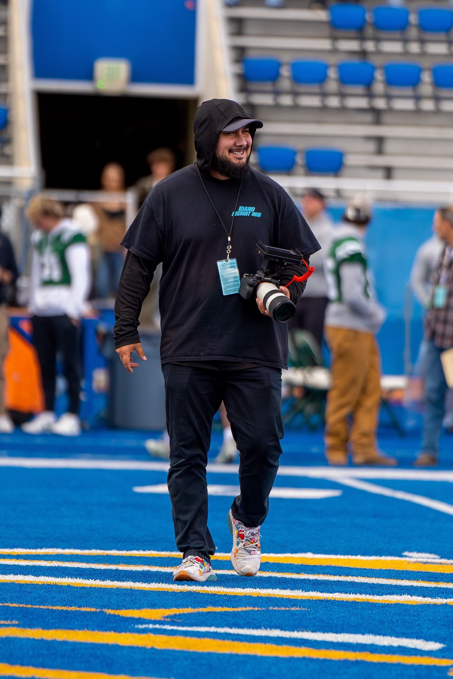 A photographer in dark clothing and beanie walks across a bright blue sports field with a camera in hand.