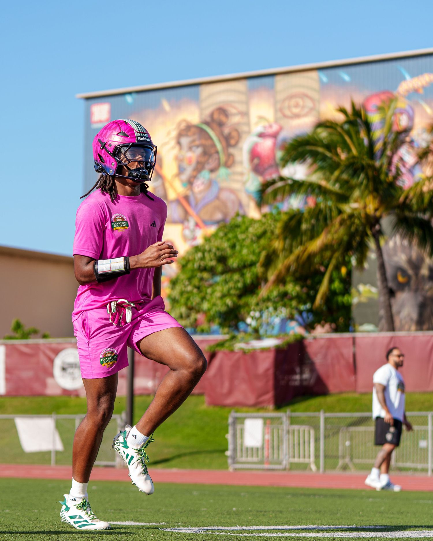 Athlete in bright pink athletic wear and helmet runs on sports field with palm trees and colorful mural in background.