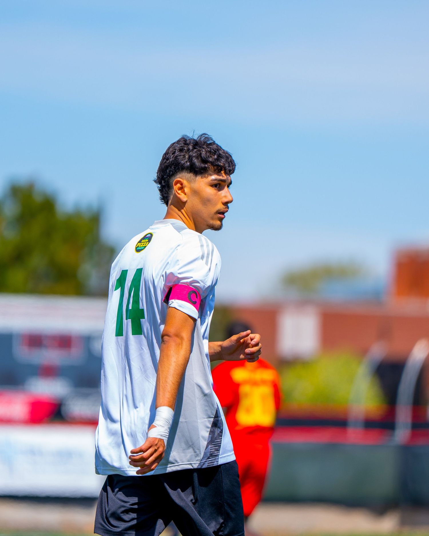 A soccer player in a white jersey with number 14 watches the action during an outdoor match on a sunny day.